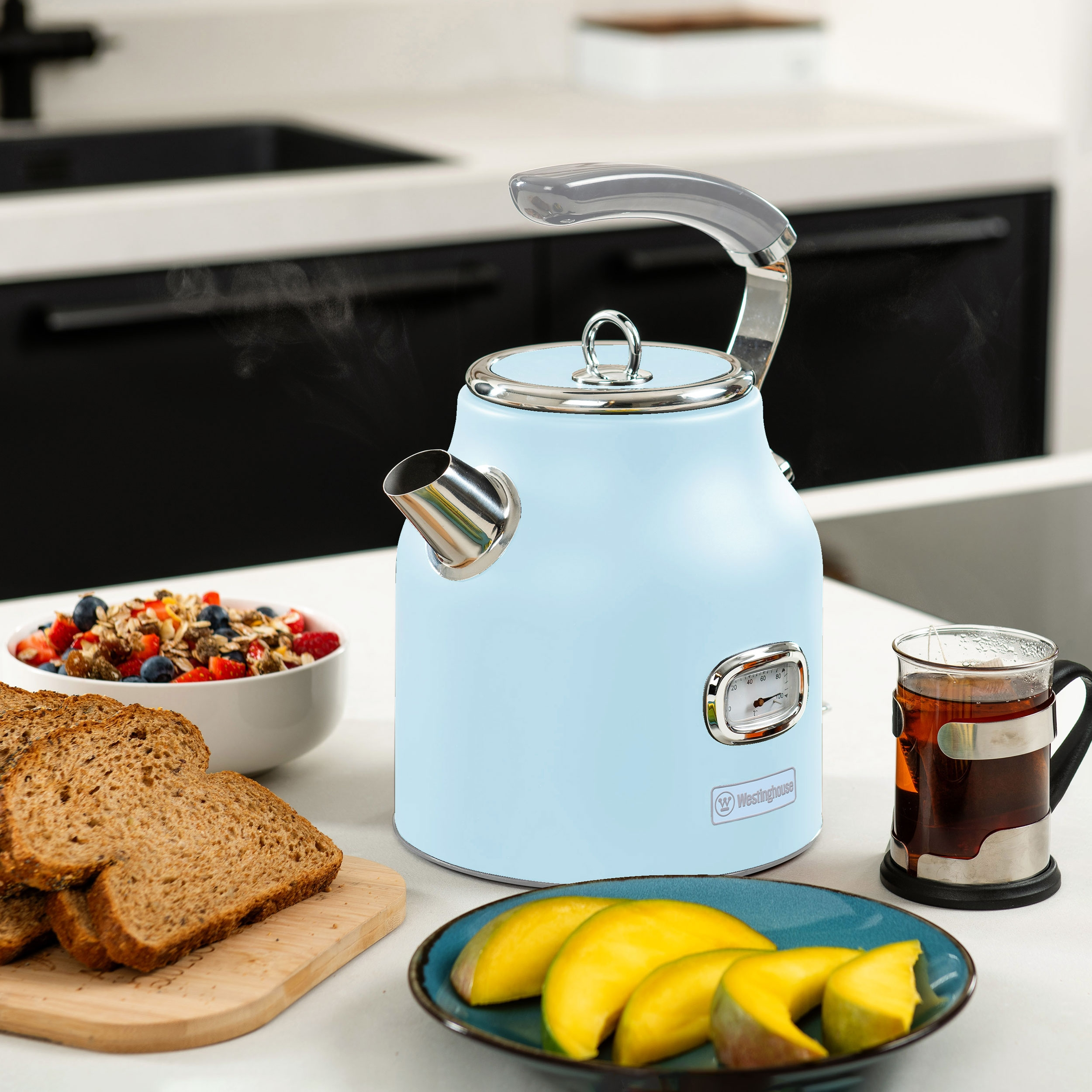 Ein hellblauer Wasserkocher mit einer Tasse Tee, Brot und Obst auf einer weißen Arbeitsplatte.