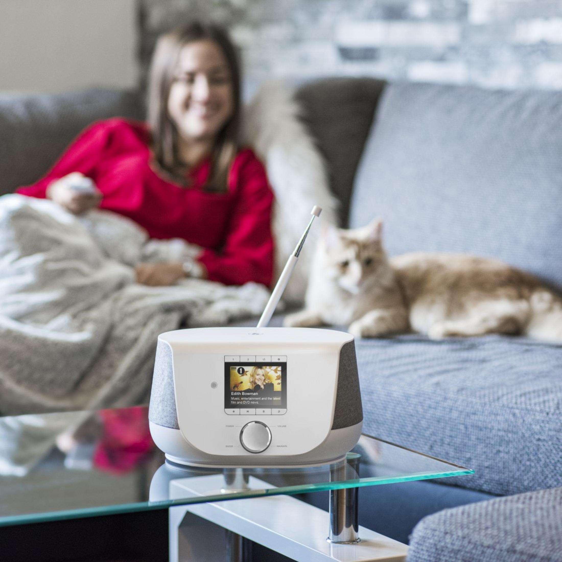 Kobieta na szarej sofie z radiem i kotem. Kobieta ma na sobie czerwień. Kot jest kremowy.