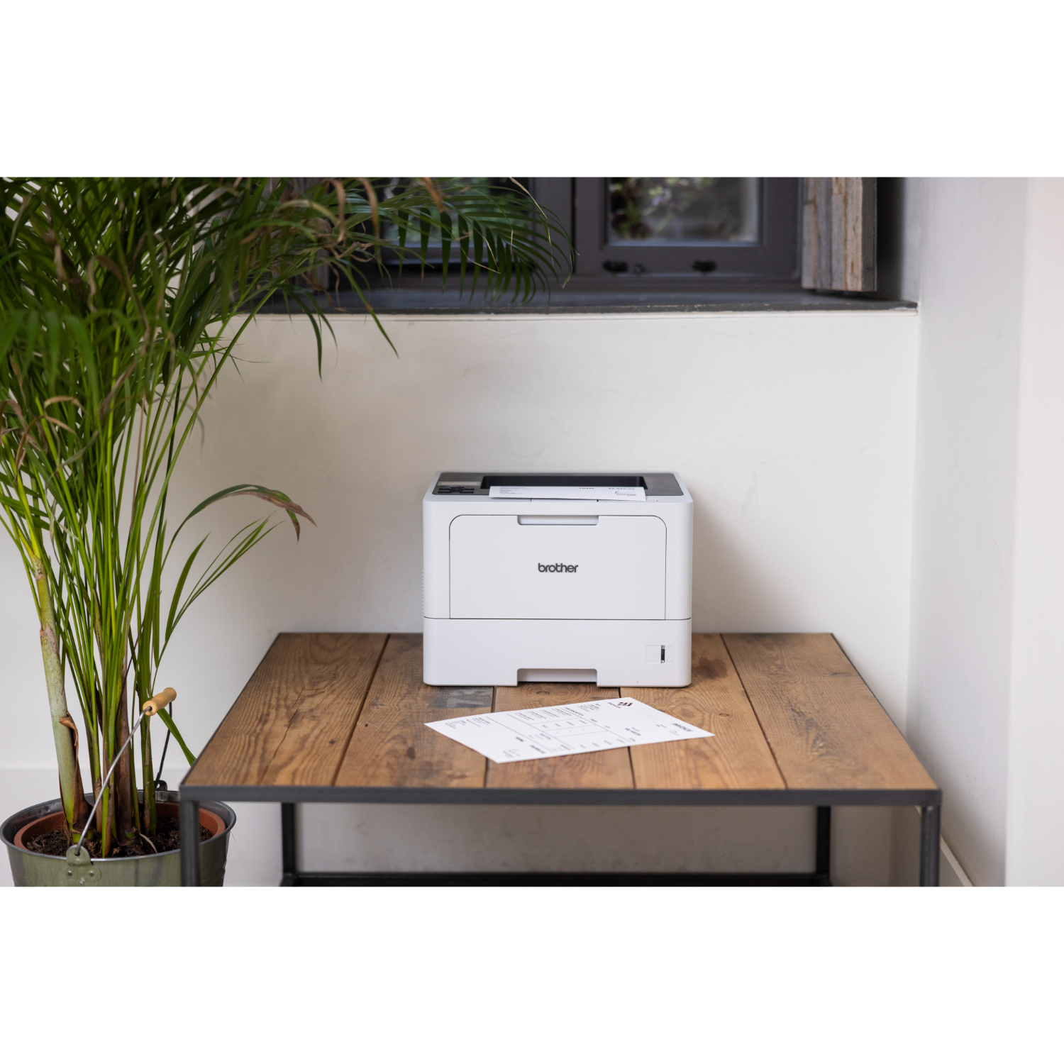 Biała drukarka na drewnianym stole z rośliną i oknem w tle.