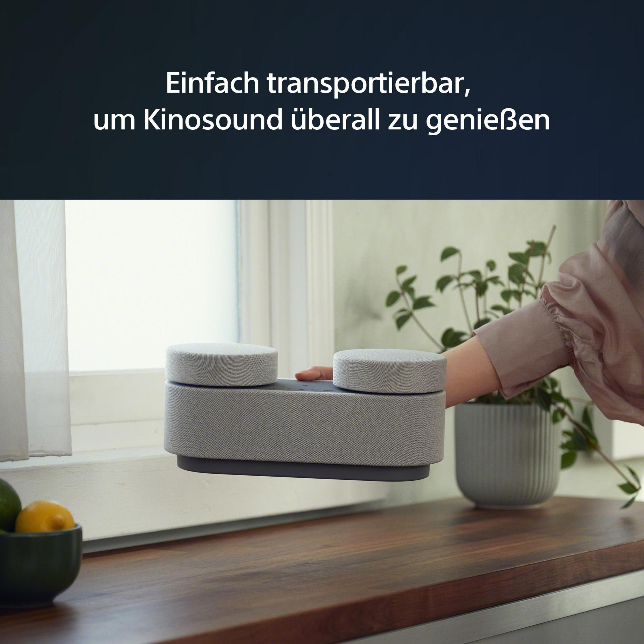 Person hält einen grauen Lautsprecher auf einem Holztisch neben einem Fenster mit Pflanzen.