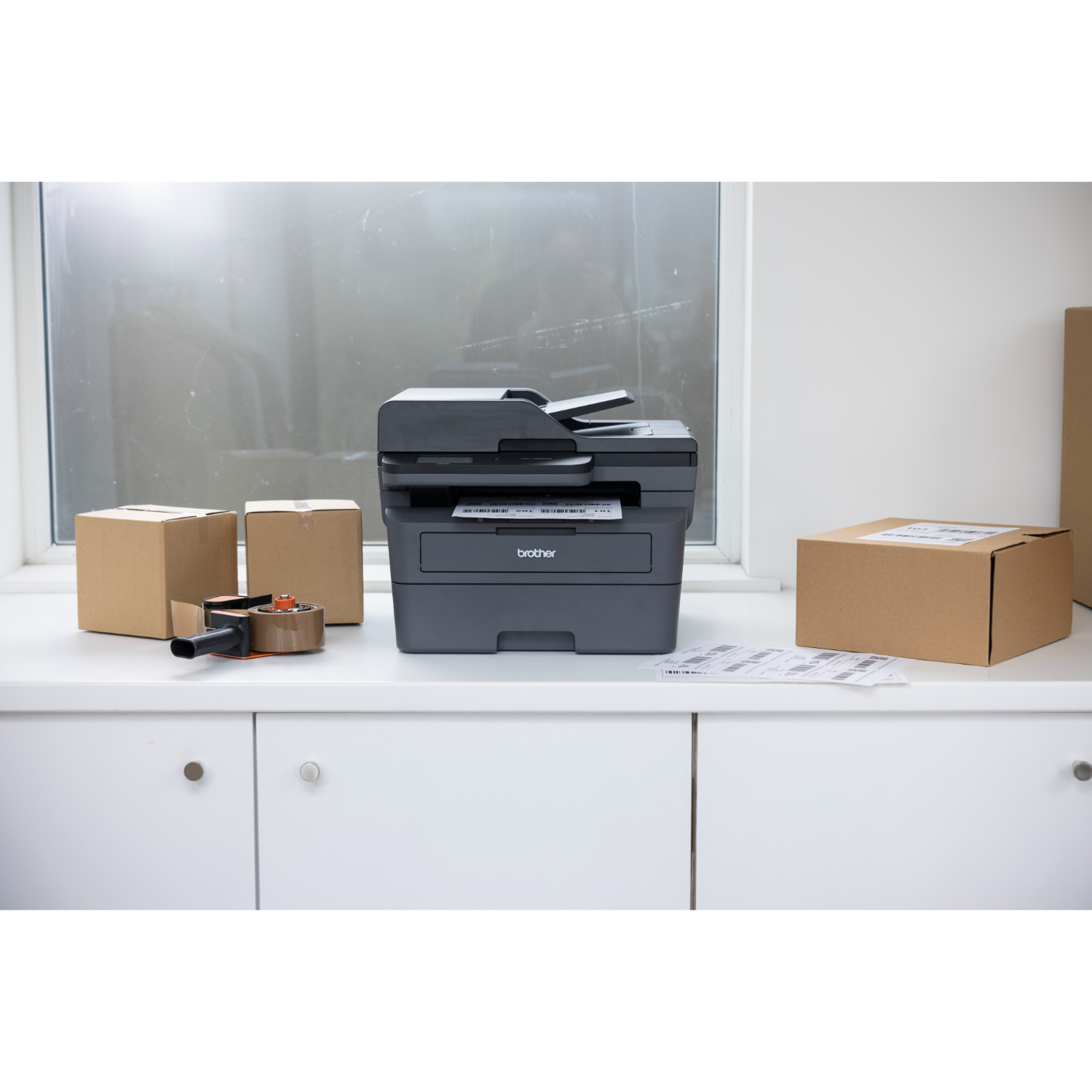 Drukarka na białym blacie z pudełkami, taśmą i papierem w pobliżu okna.