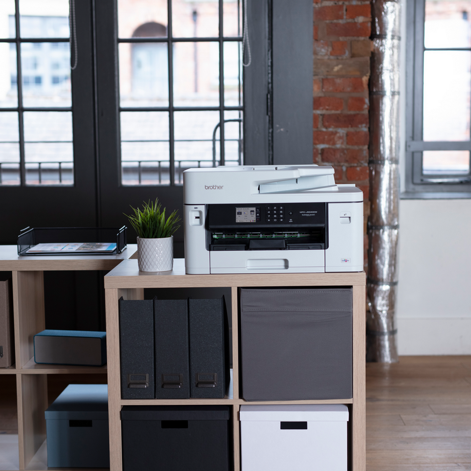 Biała drukarka na drewnianej półce w biurze. W tle znajduje się roślina, segregatory i pudełka.