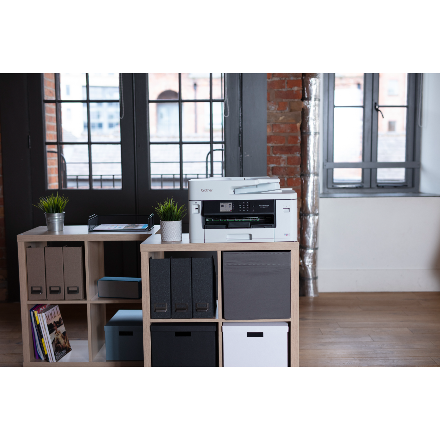 Biała drukarka na półce z kostkami wypełnionej przedmiotami. Okno i ceglana ściana w tle.