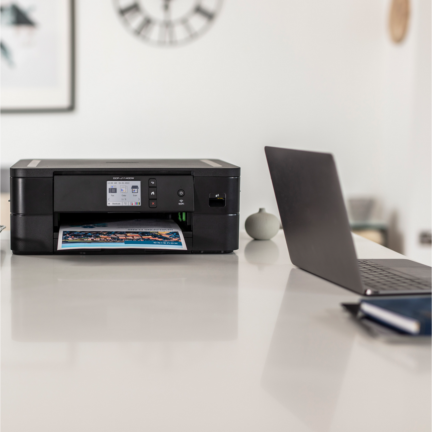 Czarna drukarka drukująca kolorowy obraz, z laptopem i małym przedmiotem dekoracyjnym.