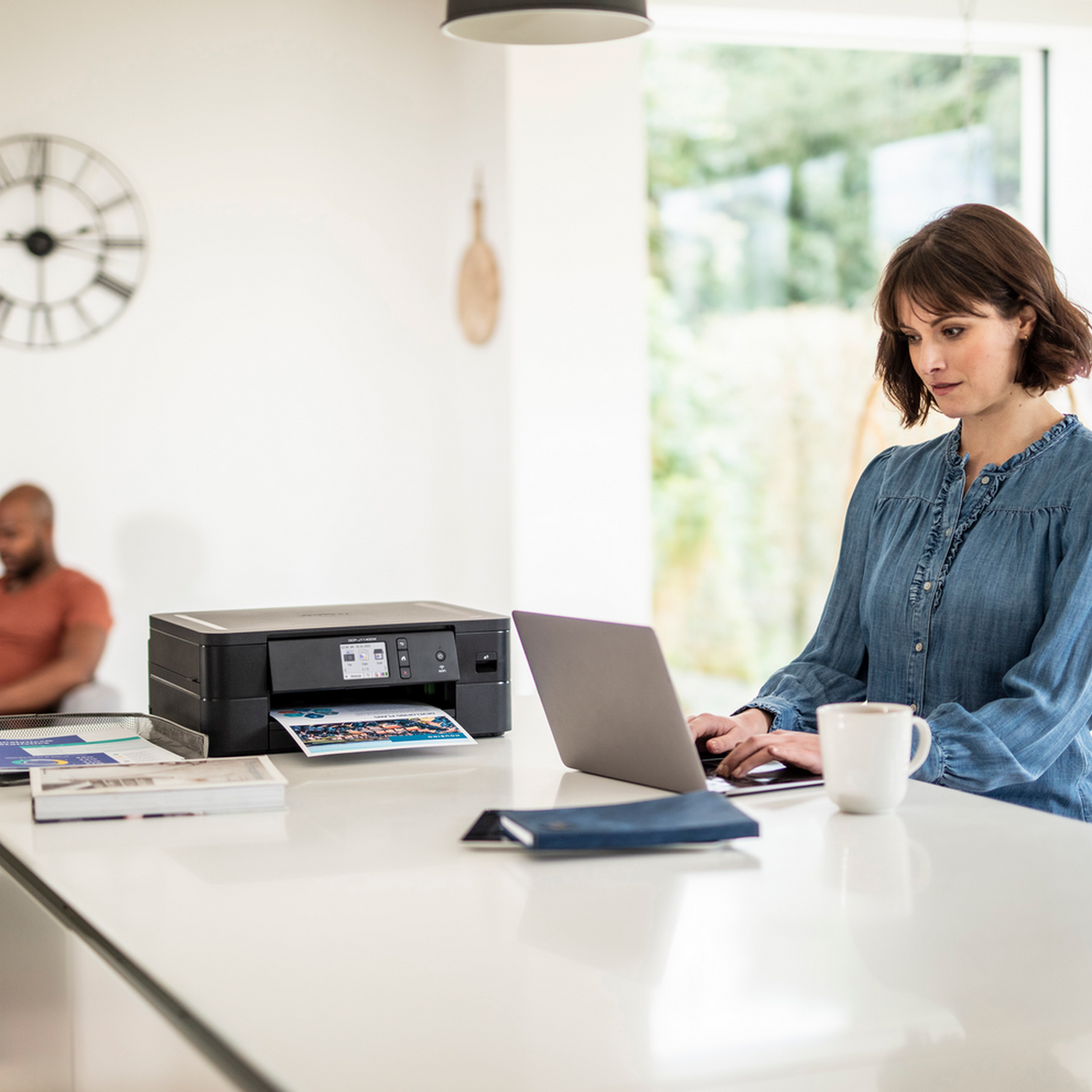 Kobieta pracuje na laptopie obok drukarki i kawy. Druga osoba w tle.