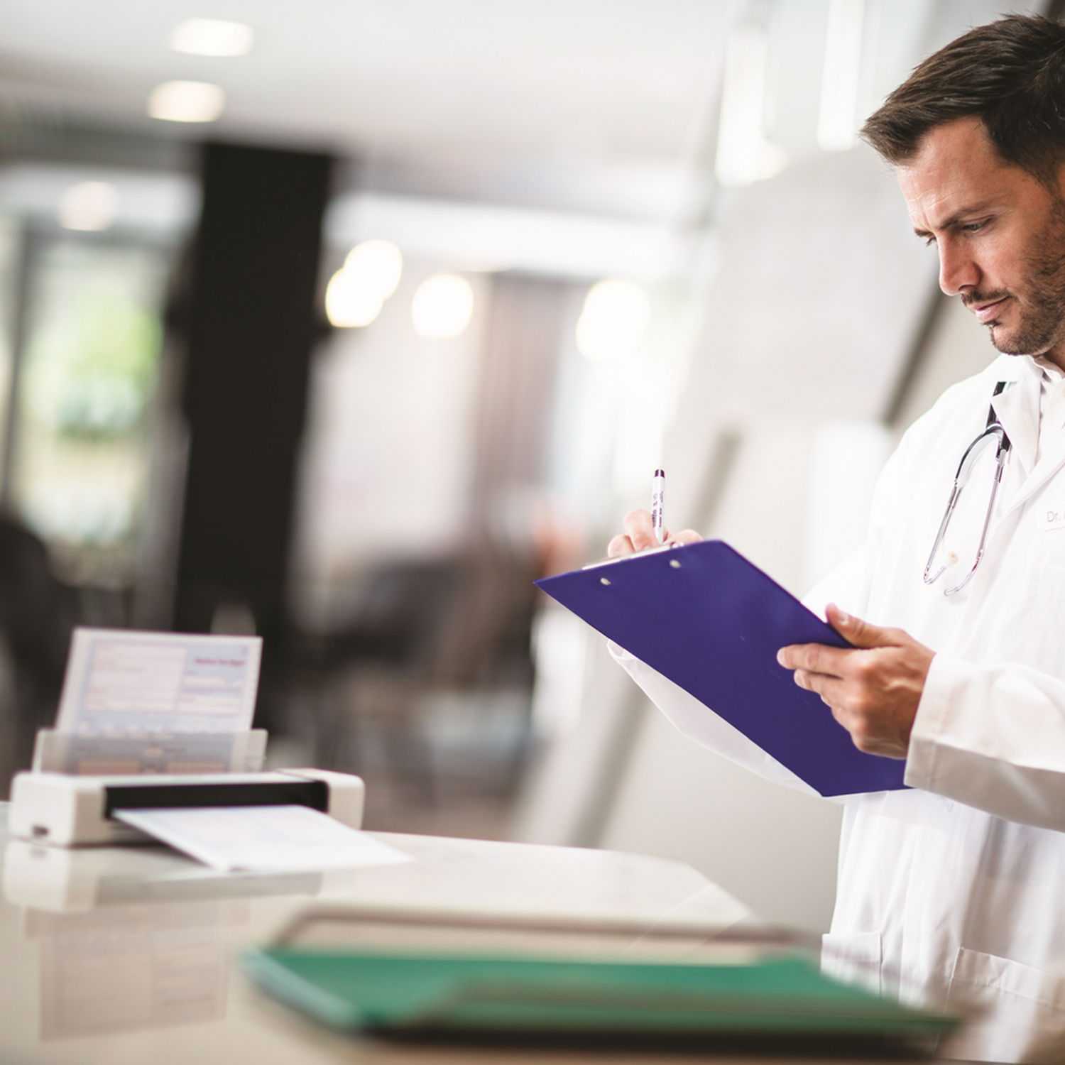 A doctor in a white coat writing on a clipboard. A printer and papers are on a counter.