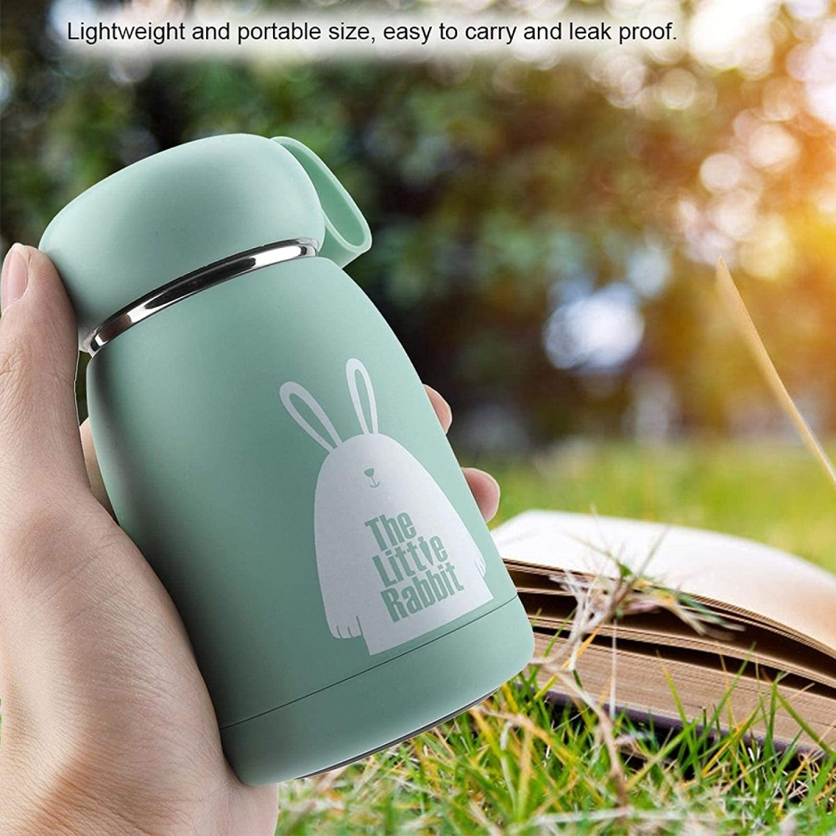Eine grüne Thermoskanne mit Hasen-Design, gehalten von einer Hand, im Hintergrund ein offenes Buch.