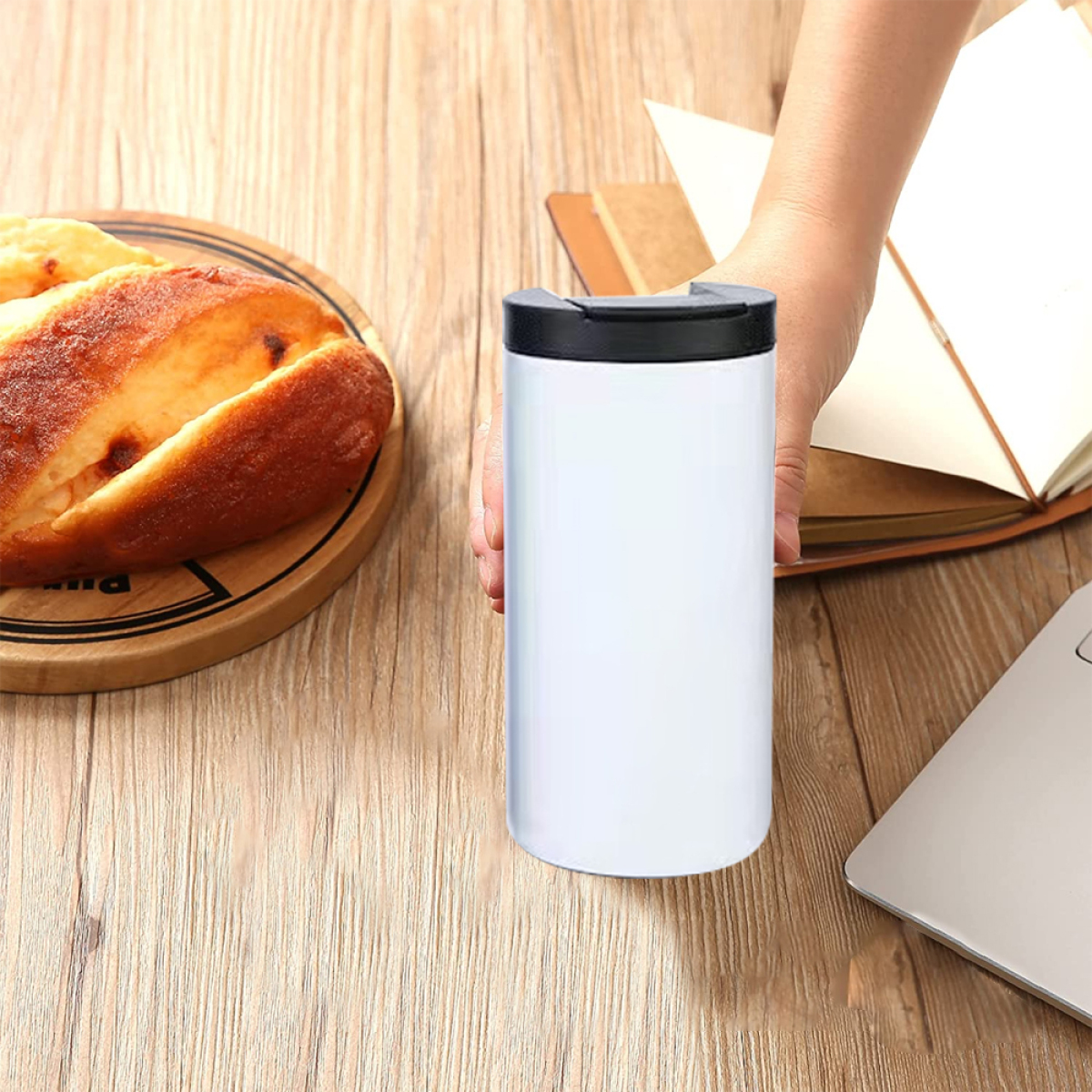 Weißer Becher in einer Hand, Holzoberfläche, Buch und Laptop.