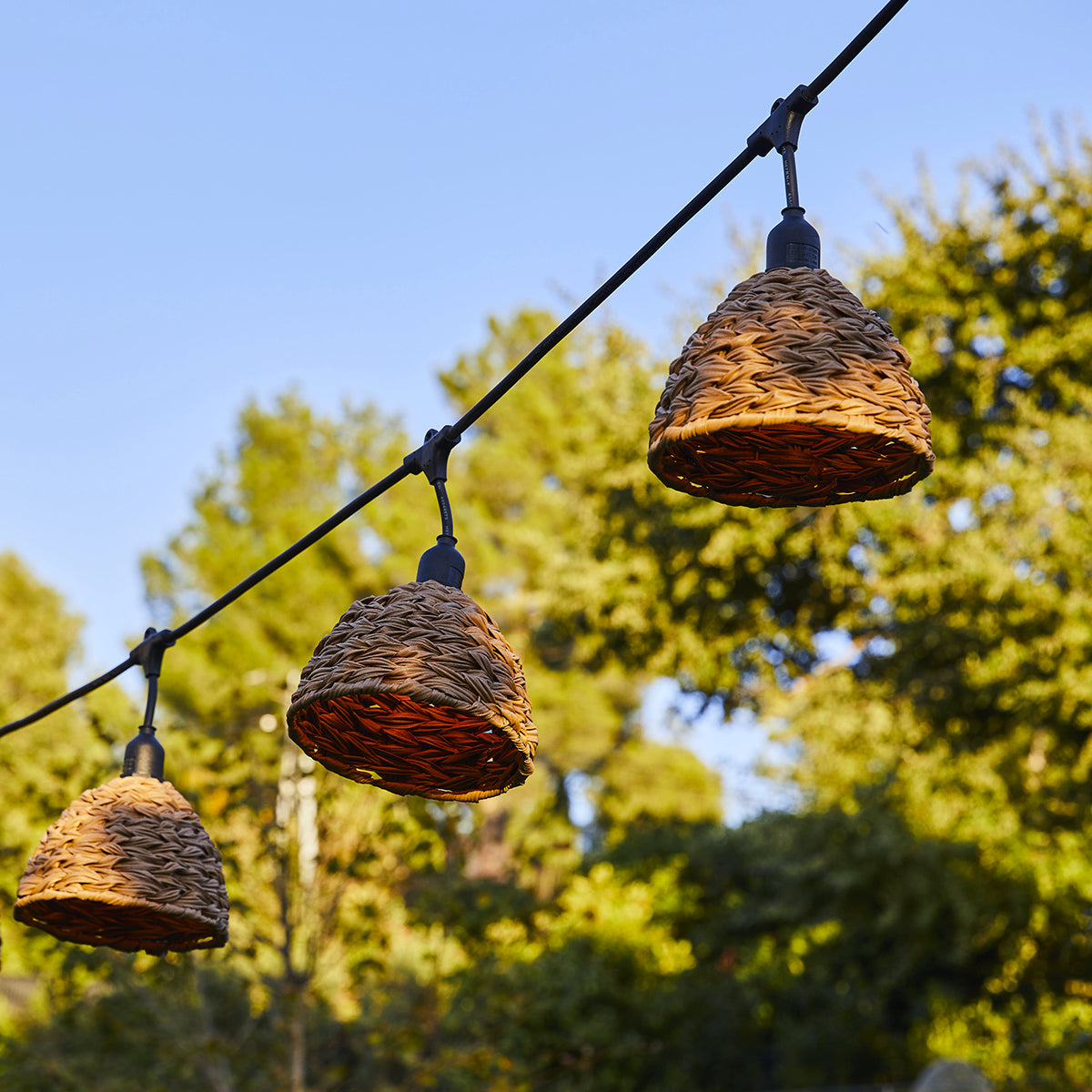Drei braune gewebte Lampen hängen an einem schwarzen Draht. Hintergrund ist blauer Himmel und Bäume.
