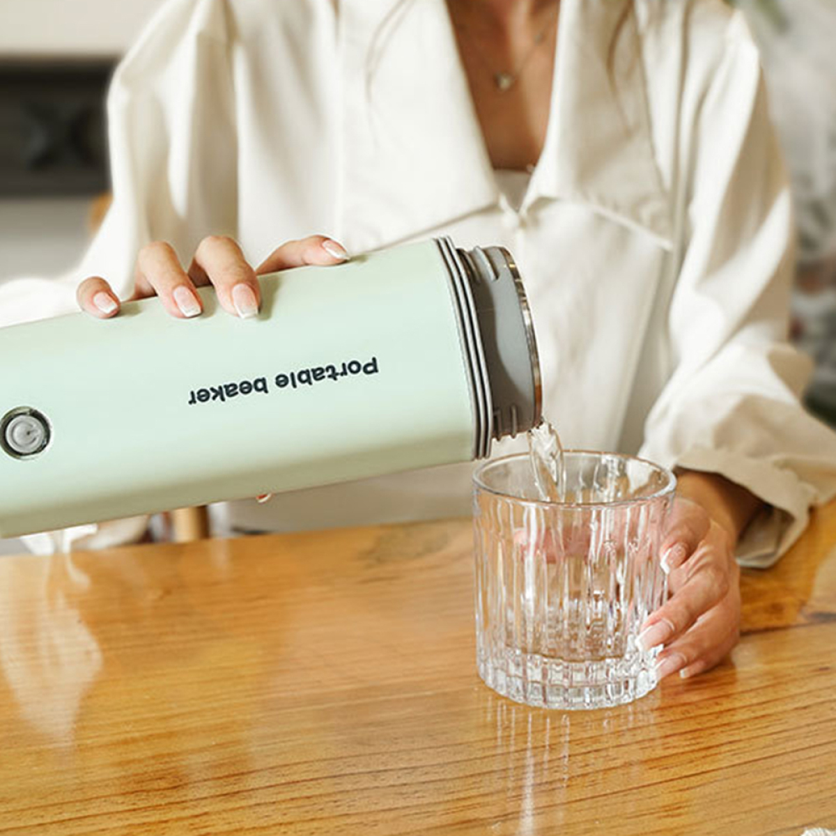 Eine Frau gießt Wasser aus einem grünen tragbaren Becher in ein Glas auf einem Holztisch.