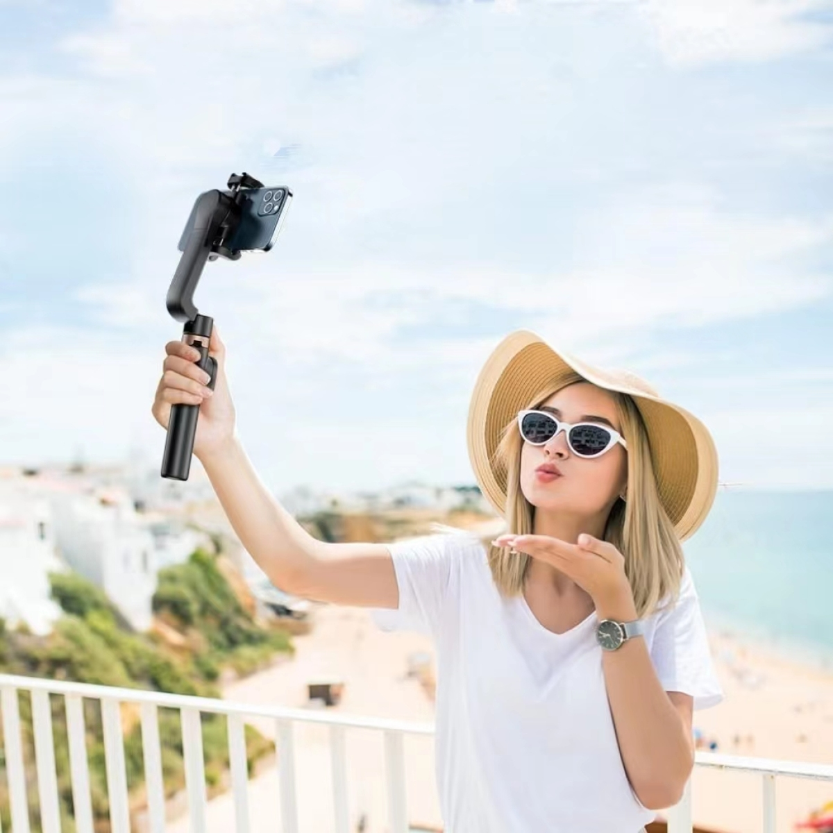 Frau macht Selfie mit Telefon auf einem Gimbal. Strandhintergrund, weißes Hemd, Hut, sonniger Tag.