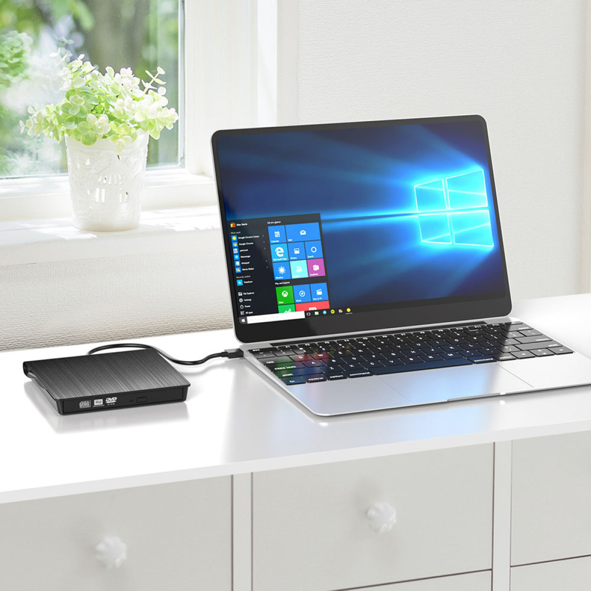 Laptop auf weißem Schreibtisch. DVD-Laufwerk angeschlossen. Fenster und Pflanze im Hintergrund. Windows-Oberfläche.