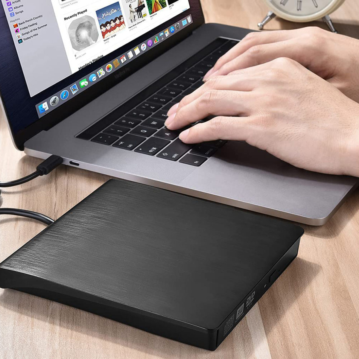 Hände tippen auf einem Laptop, mit einem schwarzen externen CD/DVD-Laufwerk verbunden. Das Laufwerk steht auf einem Holzschreibtisch.