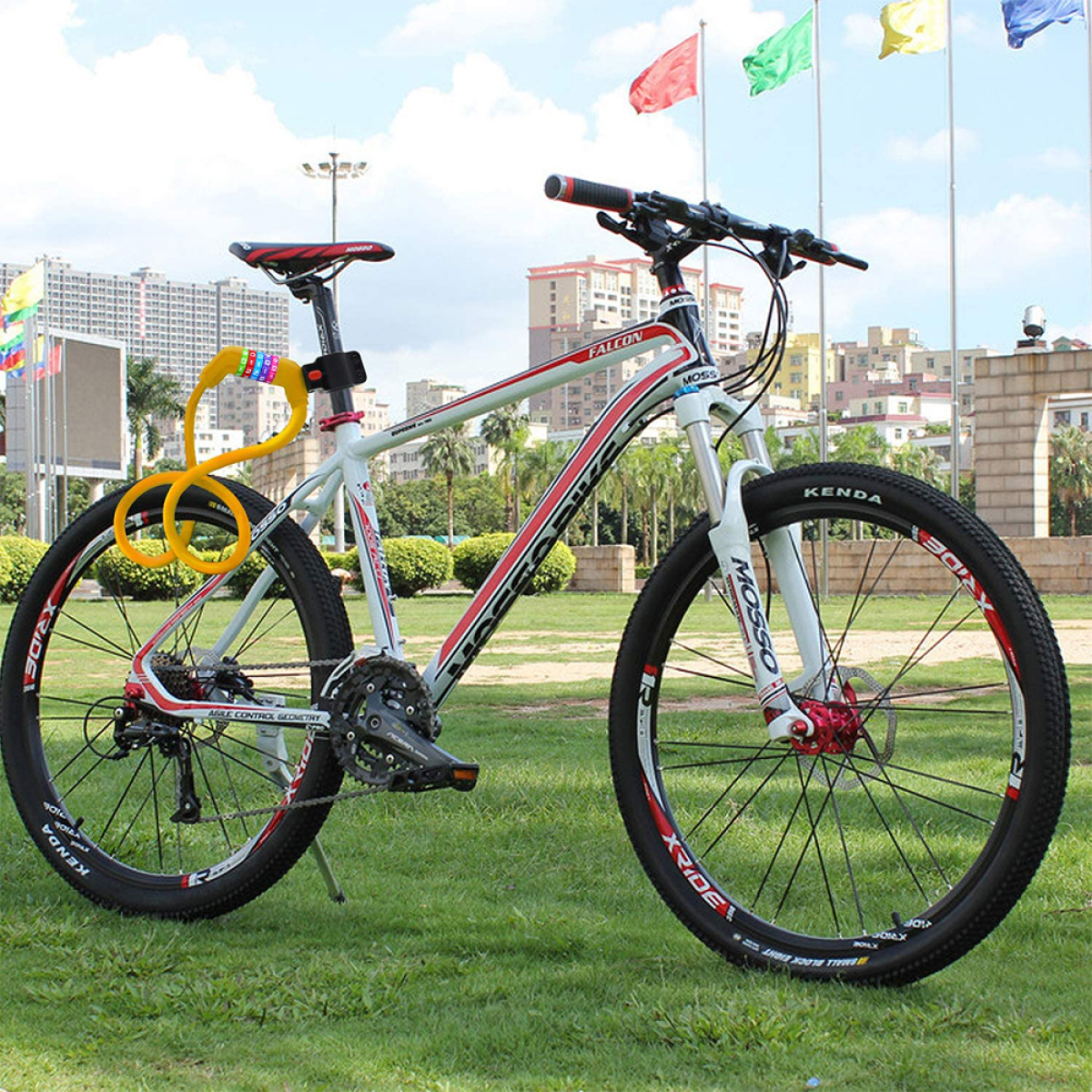 Ein weiß-rotes Fahrrad steht auf grünem Gras. Ein gelbes Schloss sichert das Fahrrad. Im Hintergrund sind Fahnen und Gebäude.