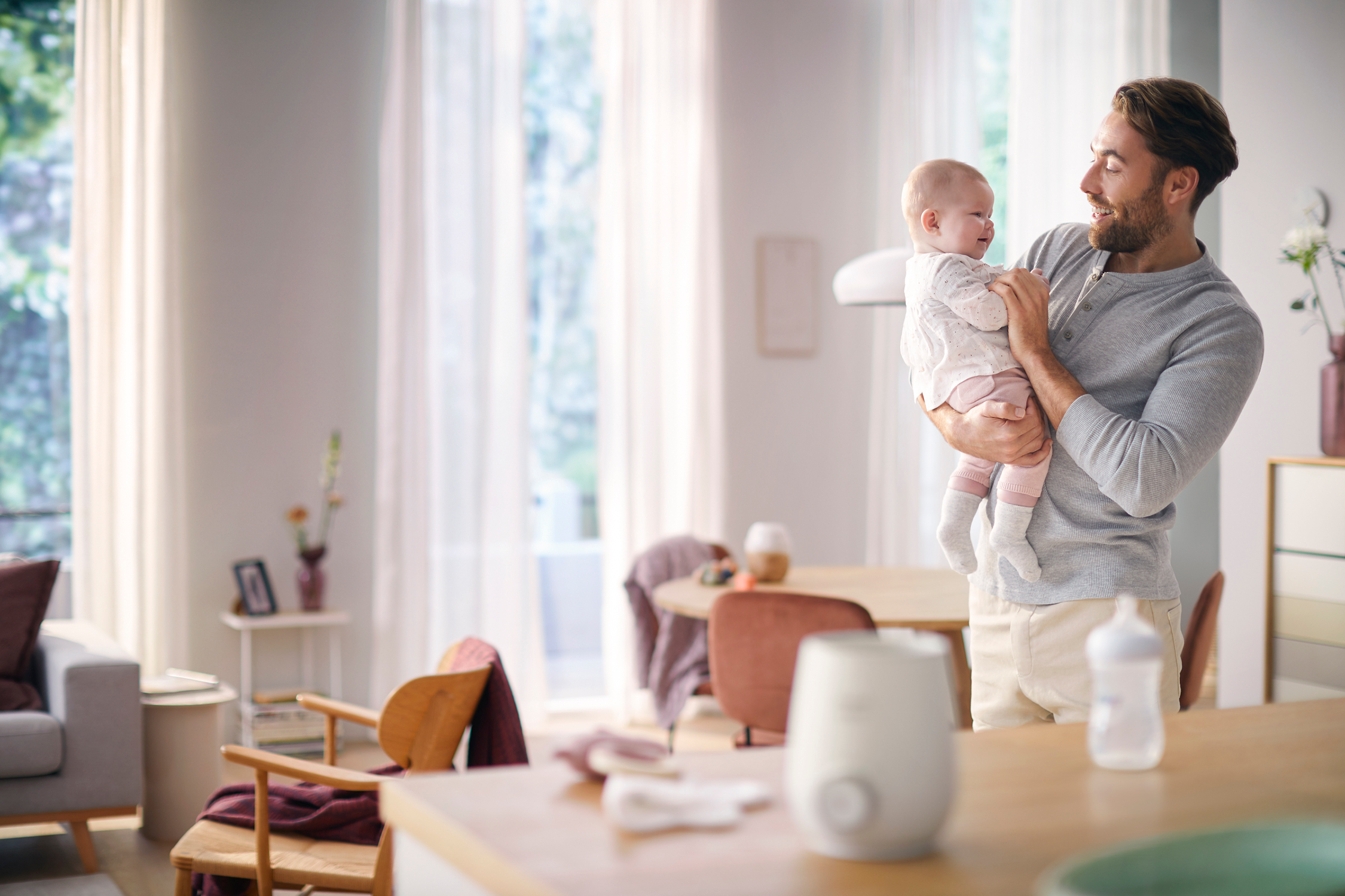 Un hombre sostiene a un bebé, sonriendo en una habitación con un calienta biberones y otros artículos.