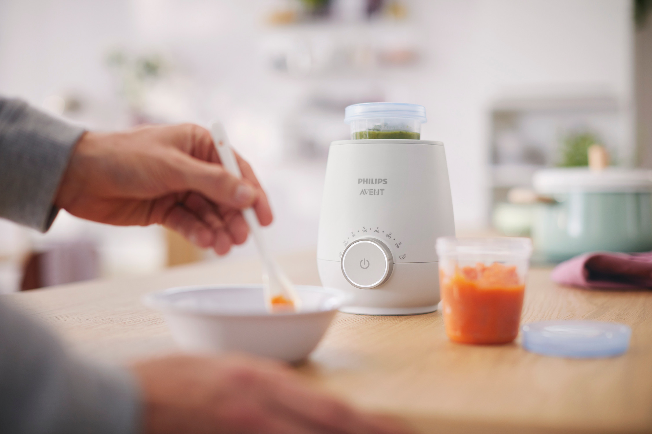Persona preparando comida para bebés en un calienta biberones blanco. Hay frascos de comida. El producto es Philips Avent.