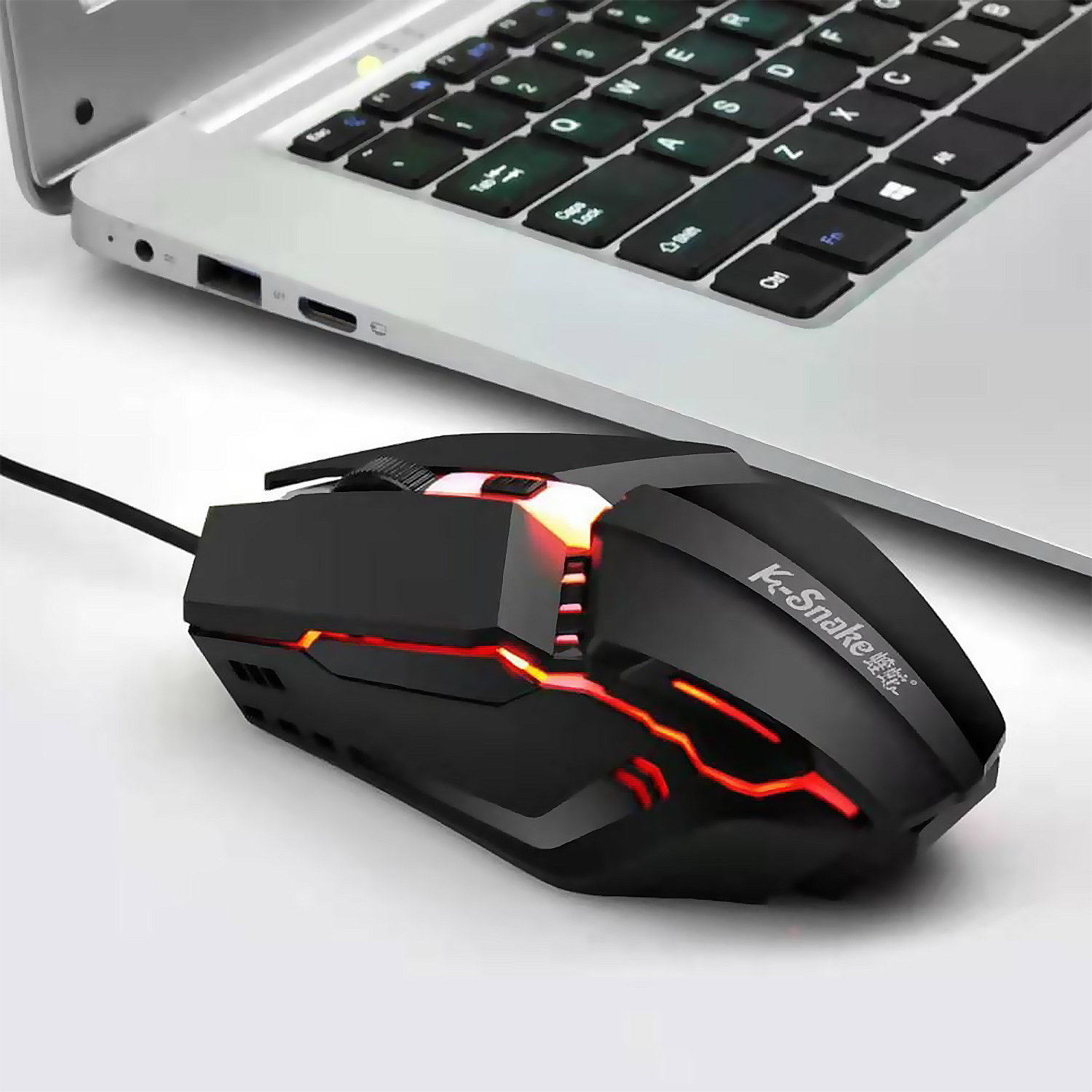 Mouse gaming nero con luci rosse, accanto a un laptop su una superficie bianca.