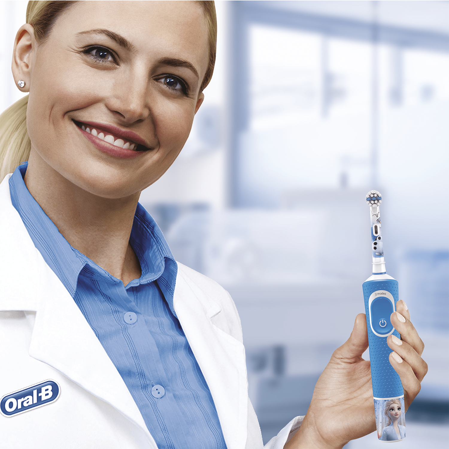 Mujer con bata de laboratorio sosteniendo un cepillo de dientes eléctrico azul y blanco con una cabeza de Elsa. Sonriendo.