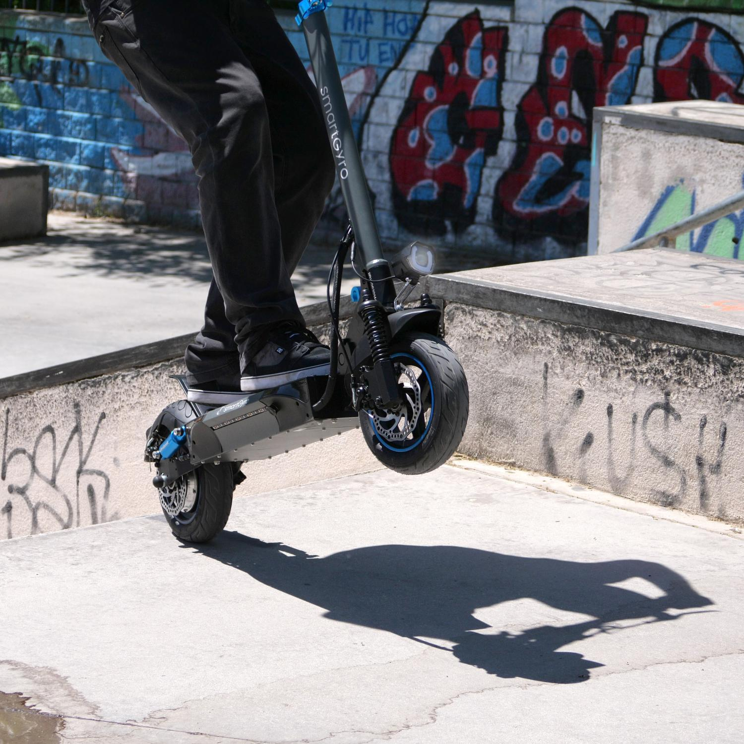 Persona montando un patinete eléctrico gris, en el aire sobre una rampa de skate park. Graffiti en el fondo.