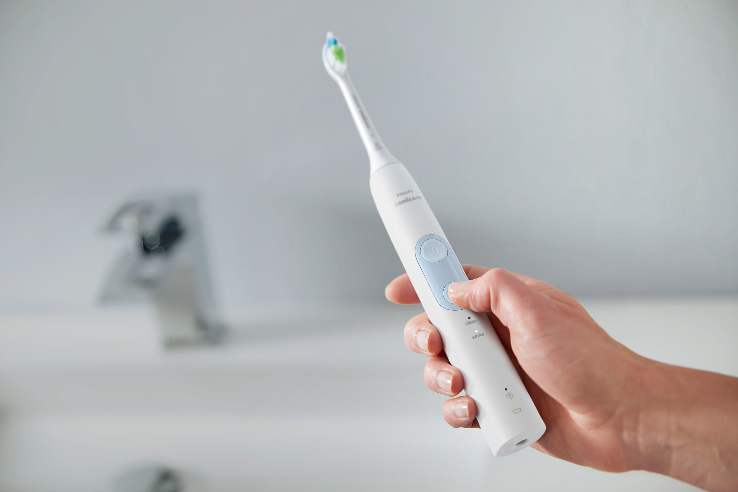 Hand hält eine weiße elektrische Zahnbürste in der Nähe eines Waschbeckens. Die Zahnbürste hat einen blauen Einschaltknopf.