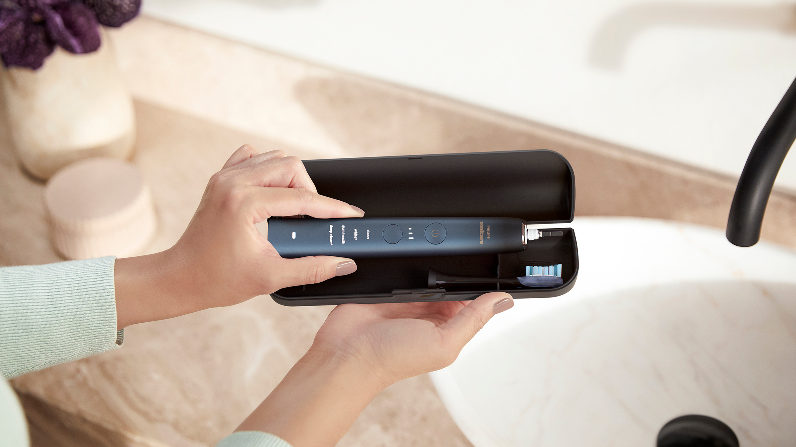 Dłonie trzymające niebieską szczoteczkę elektryczną i jej etui w pobliżu zlewu.