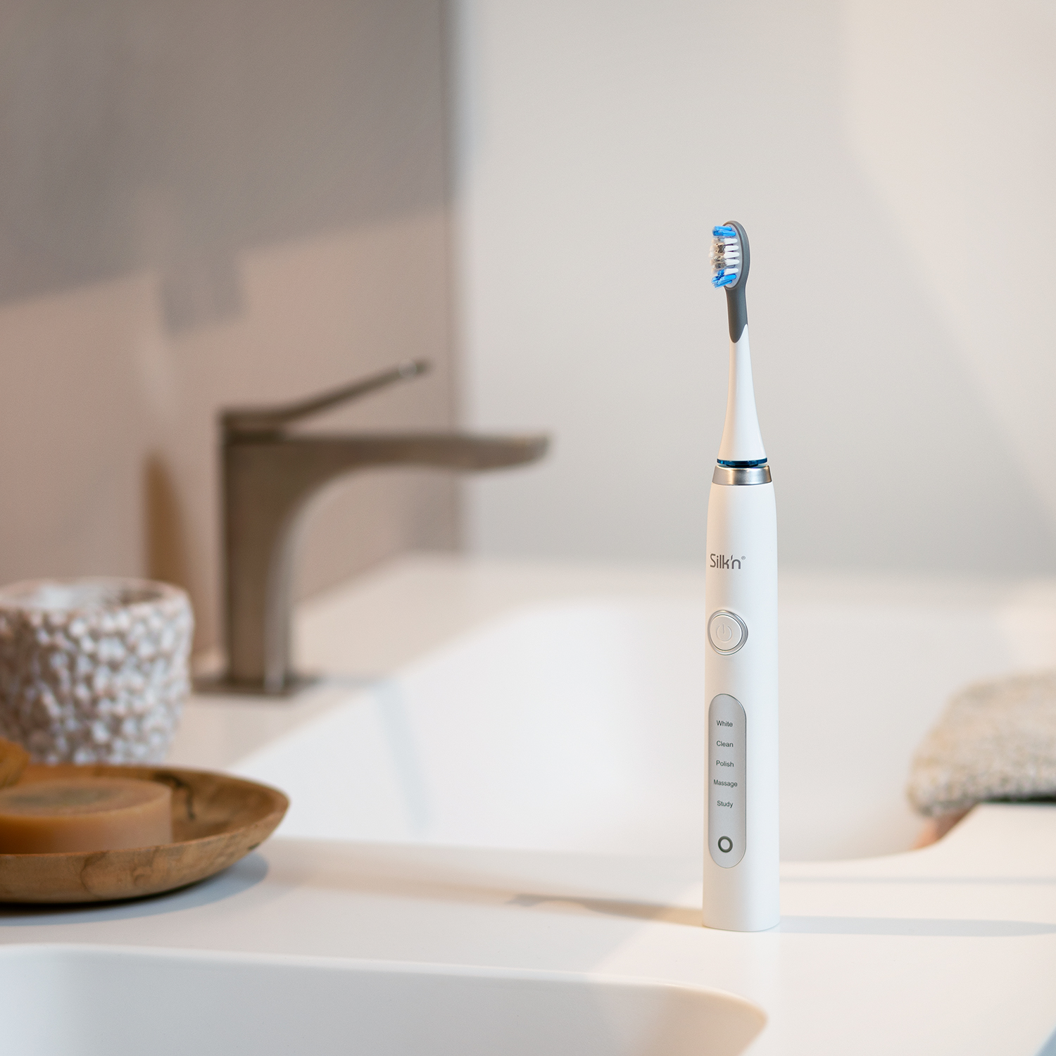 Biała elektryczna szczoteczka do zębów z niebieską główką, na białym blacie. Widoczna bateria.