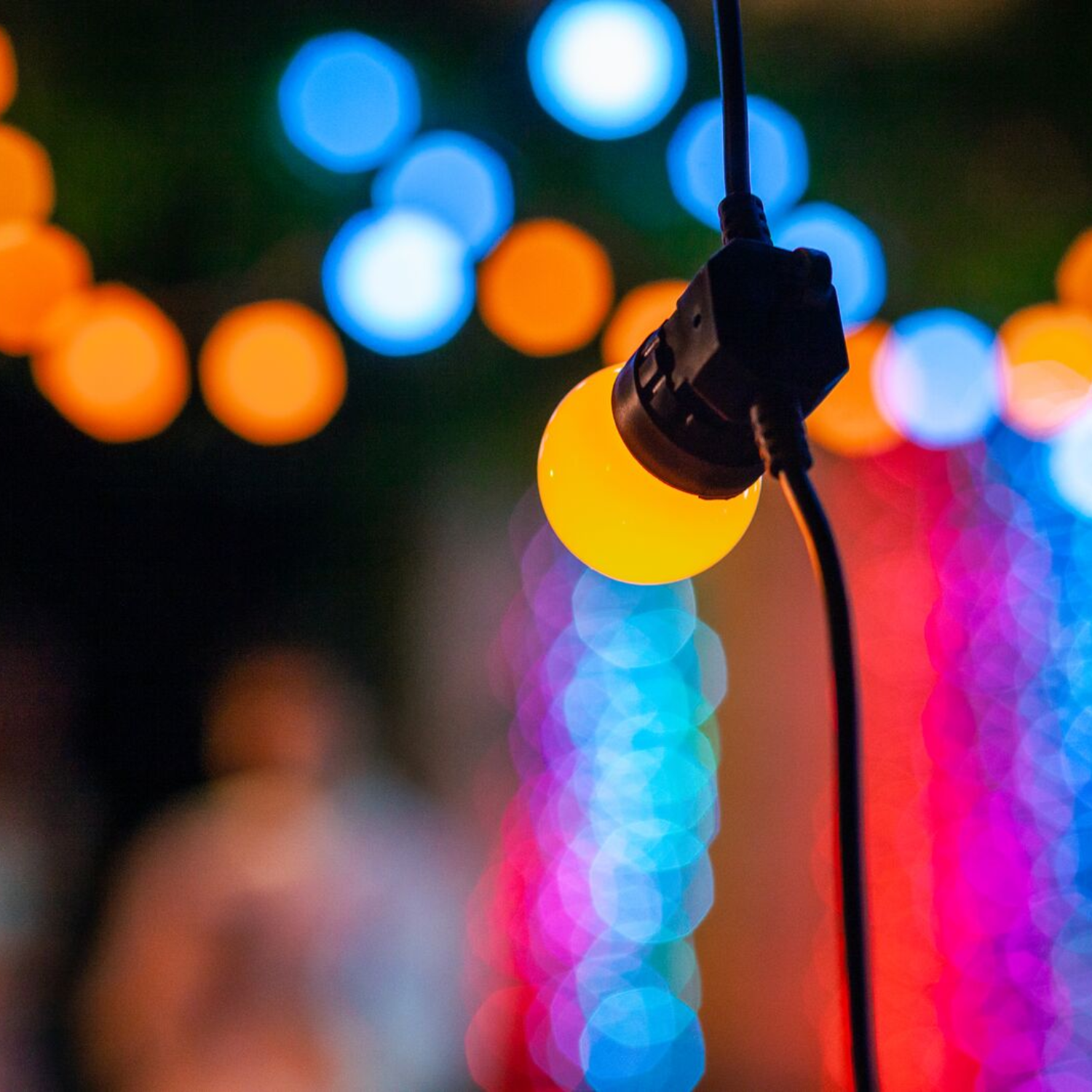 Nahaufnahme einer leuchtenden gelben Glühbirne, die an einem schwarzen Draht hängt, mit farbenfrohem Bokeh im Hintergrund.
