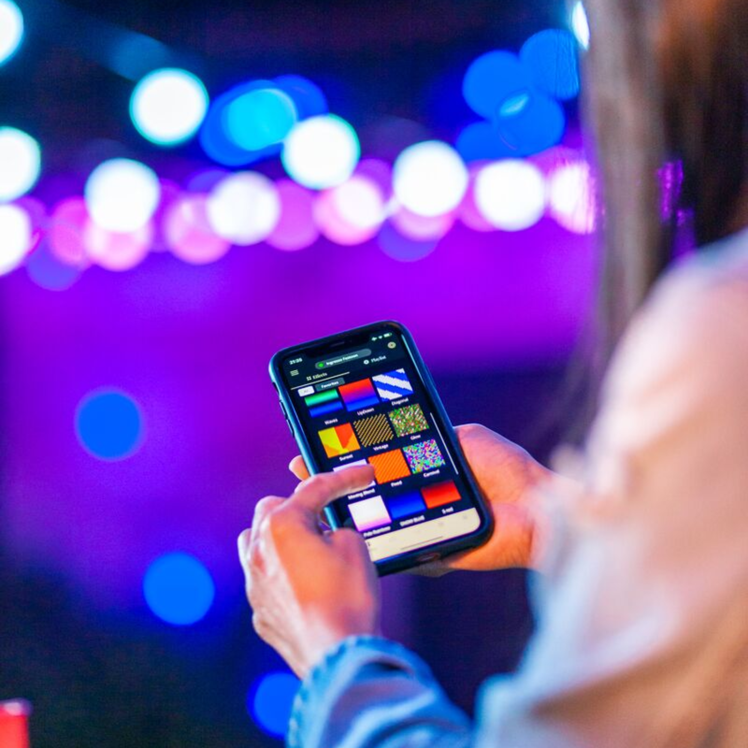 Person hält ein Handy mit Farbpallten-Oberfläche auf lila beleuchteter Bühne. Bokeh-Lichter im Hintergrund.