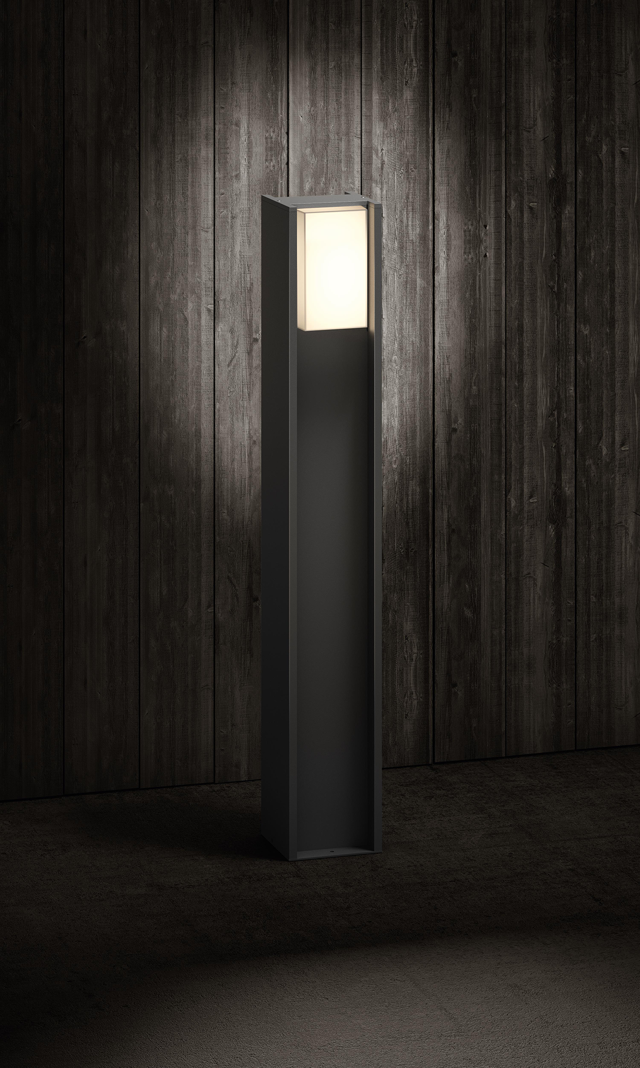 Wysoka, prostokątna, czarna lampa zewnętrzna z białym panelem świetlnym na górze. Ciemne drewniane tło.