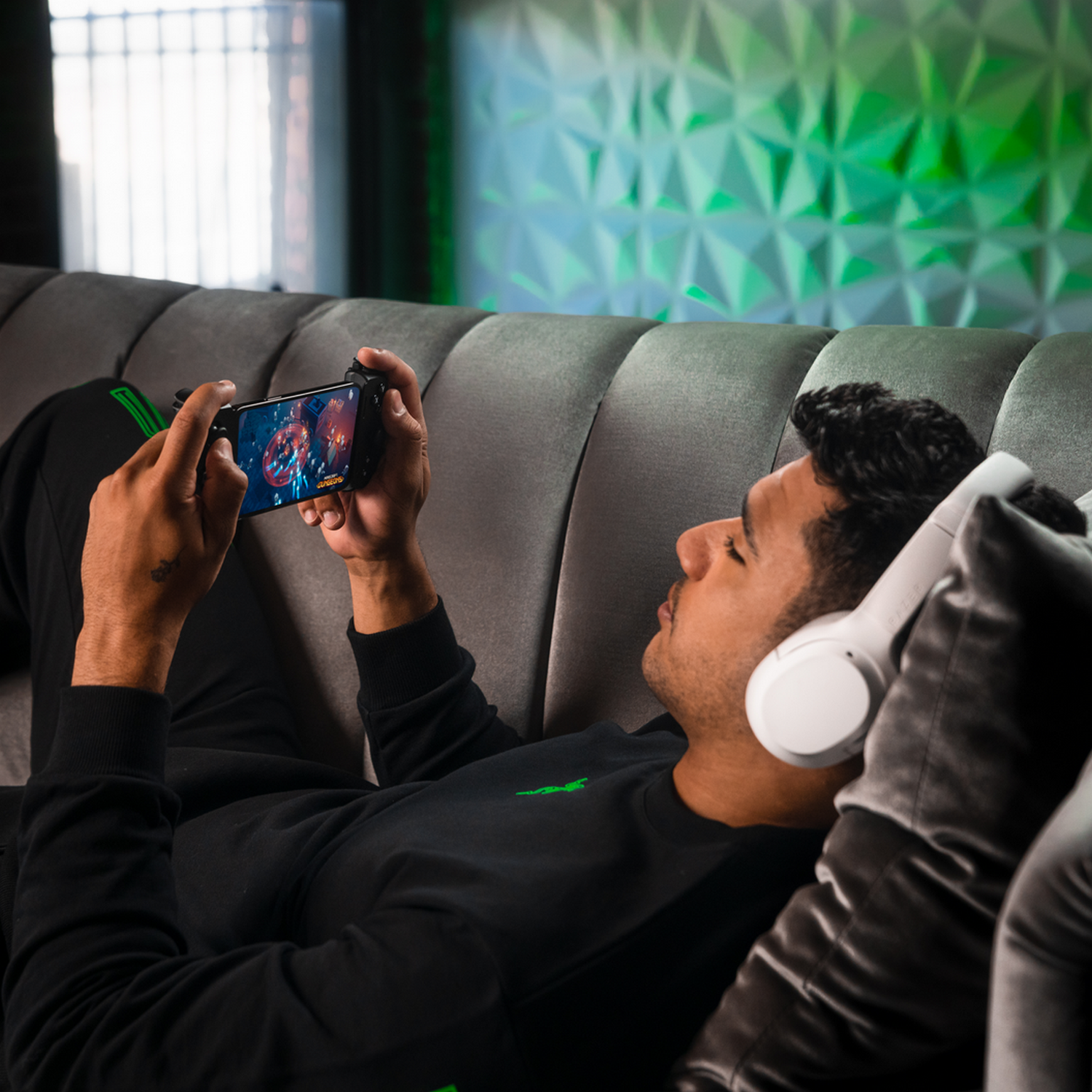 Mężczyzna leżący na kanapie gra w grę na telefonie z podłączonym kontrolerem. Ma słuchawki.
