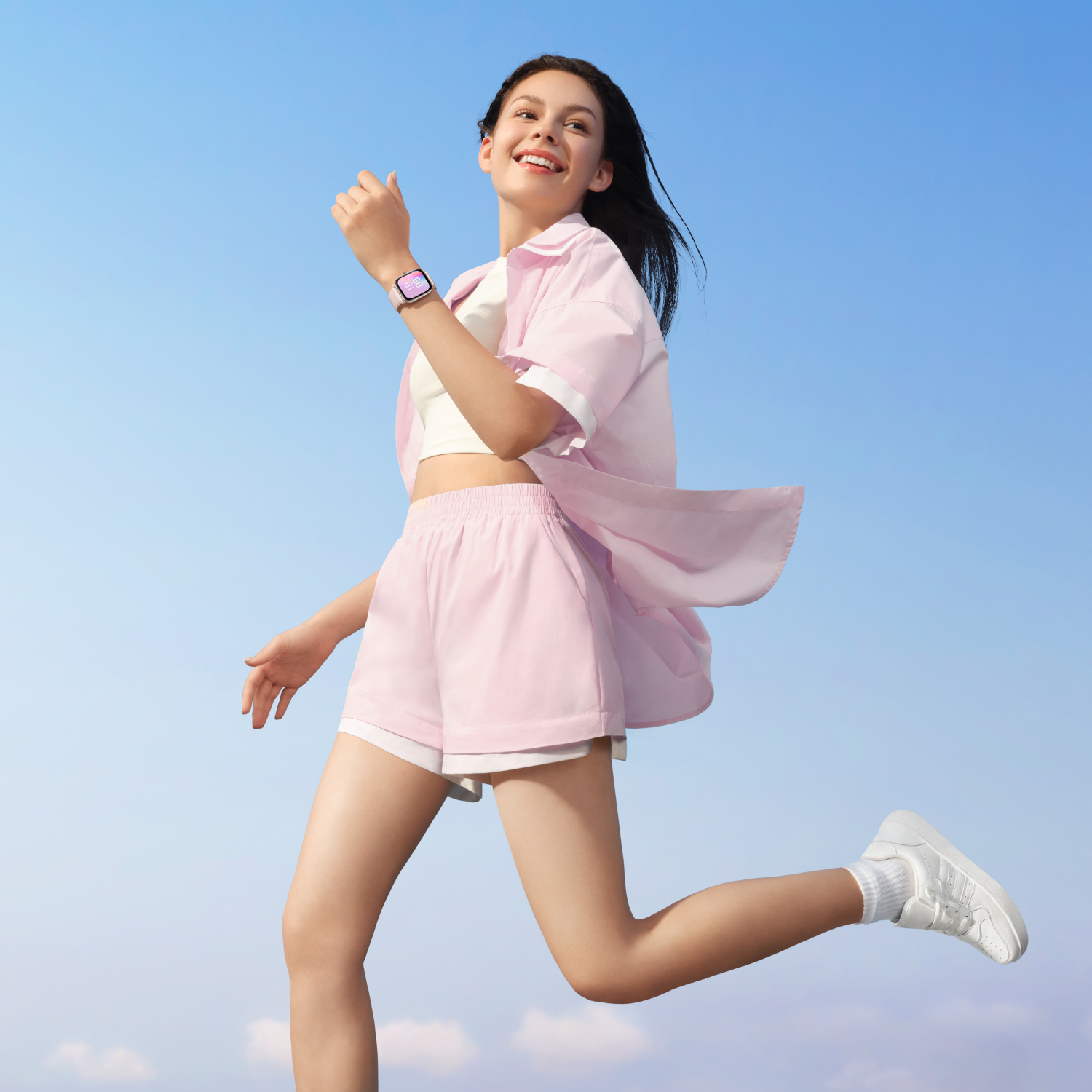 Donna che corre e sorride, indossa pantaloncini rosa, camicia e smartwatch su sfondo cielo blu.