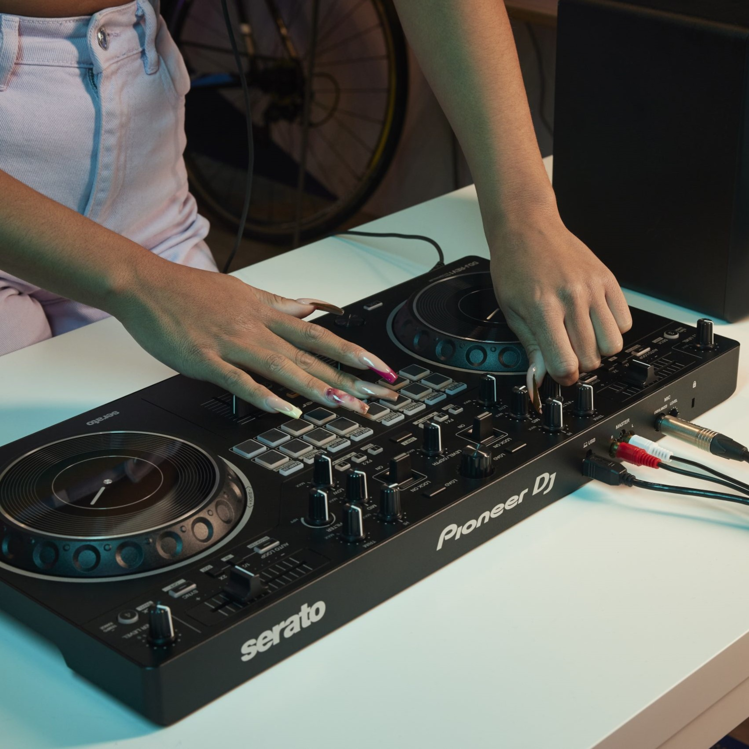 Hände, die einen DJ-Controller auf einem weißen Tisch bedienen. Kabel und ein Lautsprecher sind sichtbar.