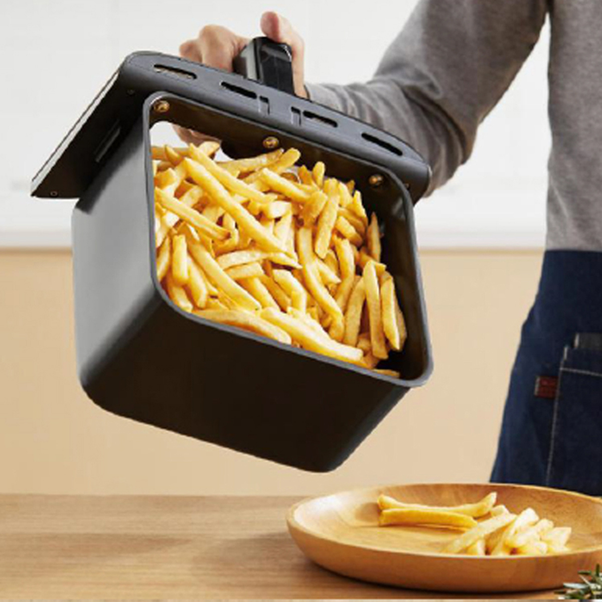 Una persona sosteniendo una cesta de freidora de aire negra llena de papas fritas doradas, con más papas fritas cerca.