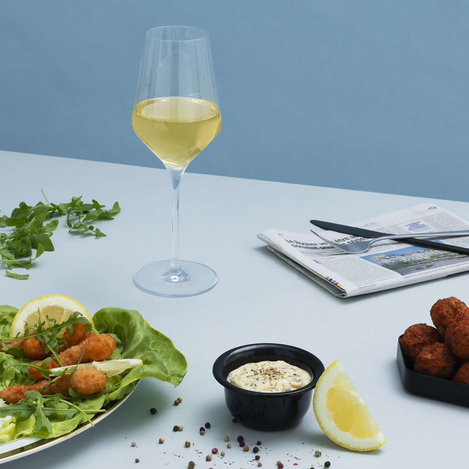 Kieliszek białego wina, jedzenie i gazeta na jasnoniebieskiej powierzchni. Jest cytryna.