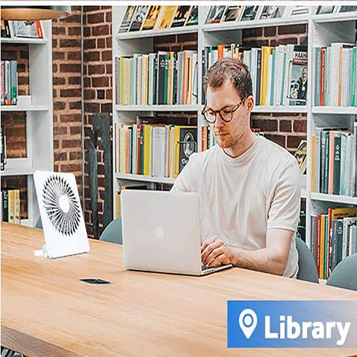Mann in einer Bibliothek, der einen Laptop benutzt, mit einem Ventilator auf dem Tisch.