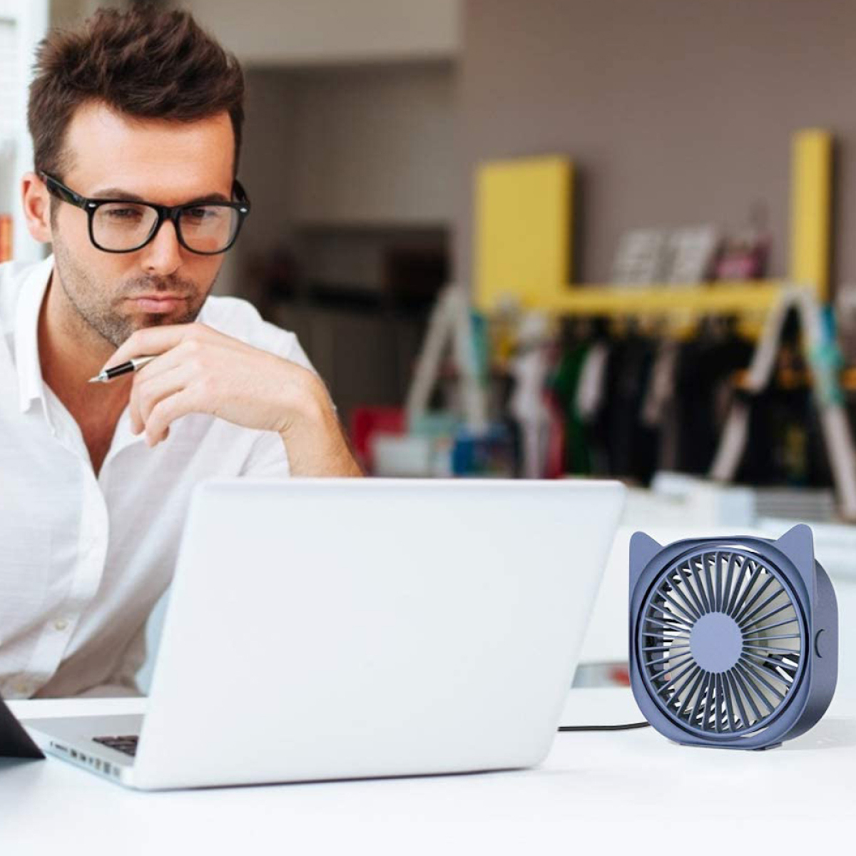 Ein Mann mit Brille schaut auf einen Laptop. Ein blauer Ventilator steht auf dem Schreibtisch. Der Hintergrund ist unscharf.