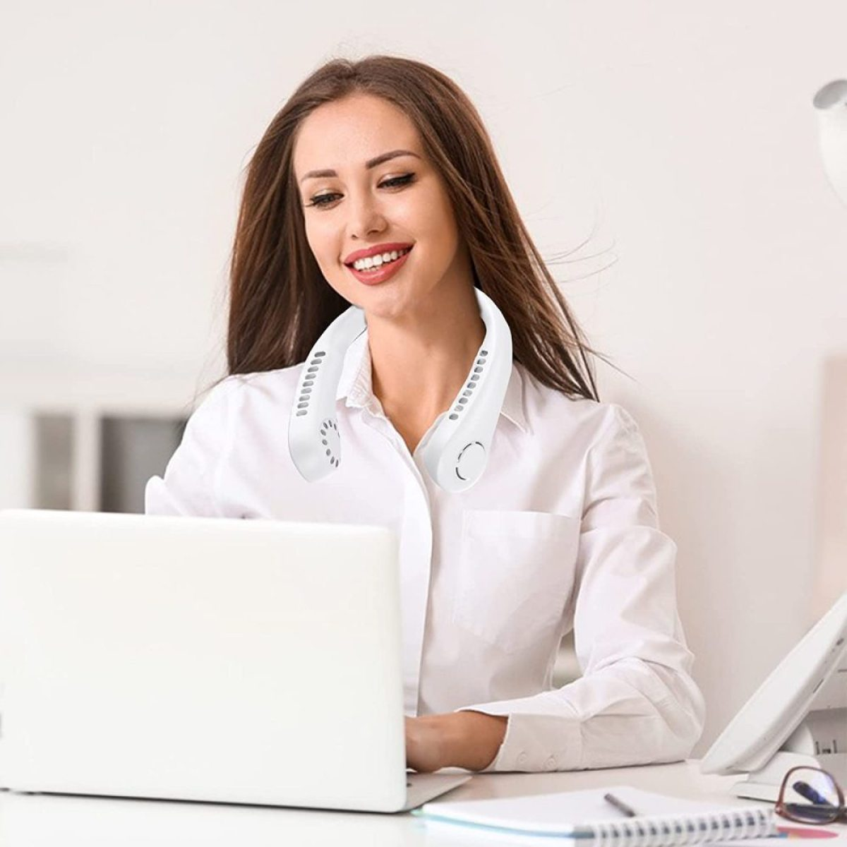 Frau in weißem Hemd, die einen weißen Nackenventilator trägt, arbeitet am Laptop.
