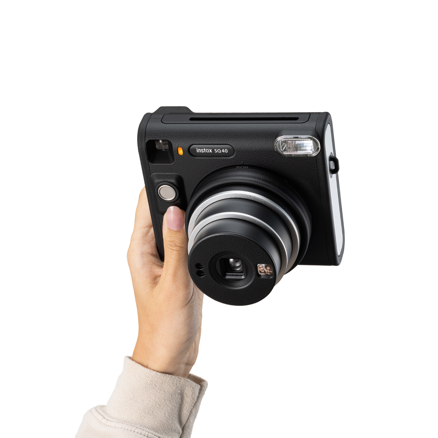 Eine schwarze Instax SQ40 Kamera wird von einer Hand gehalten, weißer Hintergrund. Objektiv und Blitz sichtbar.