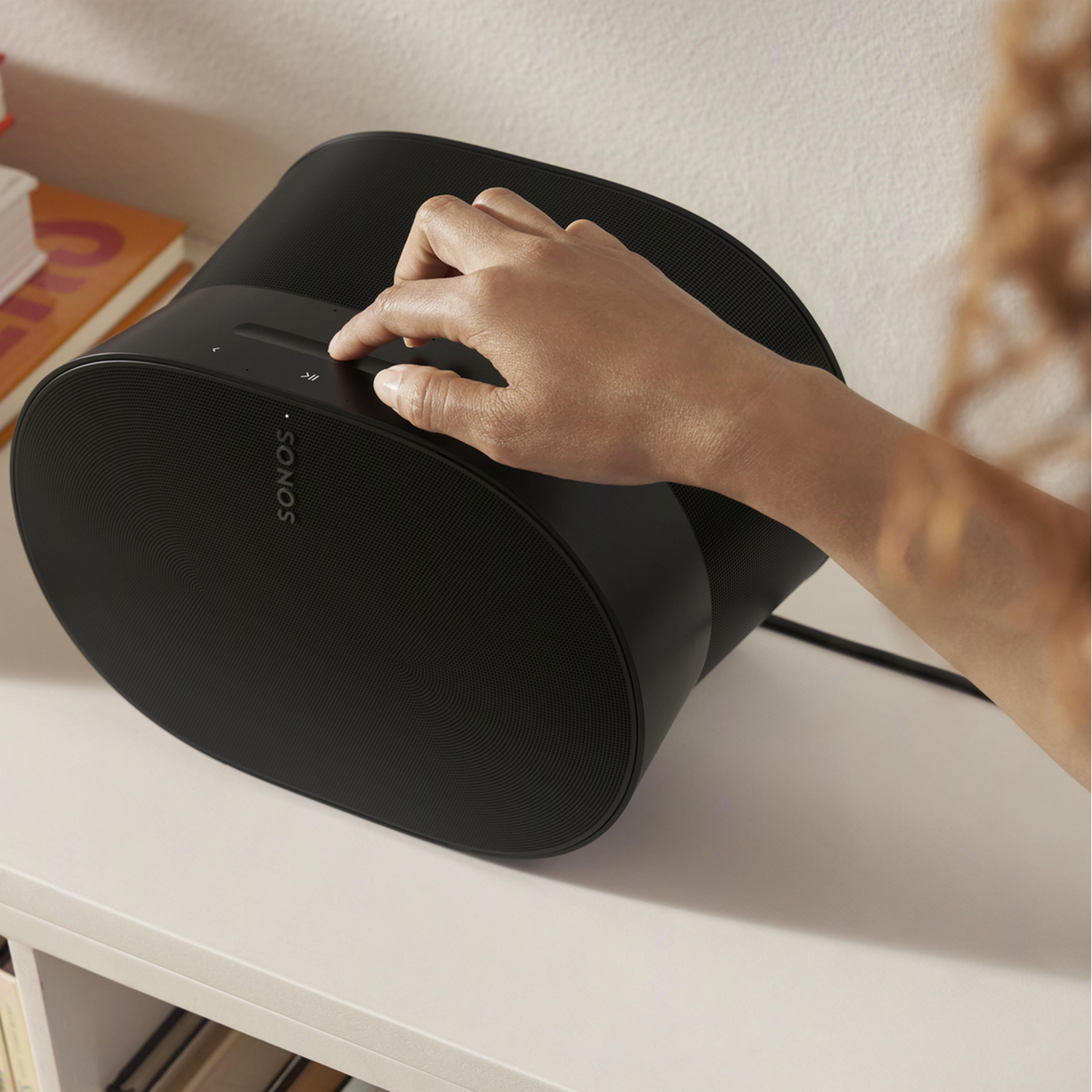 Ręka reguluje pokrętło na czarnym głośniku Sonos. Biała półka trzyma głośnik.