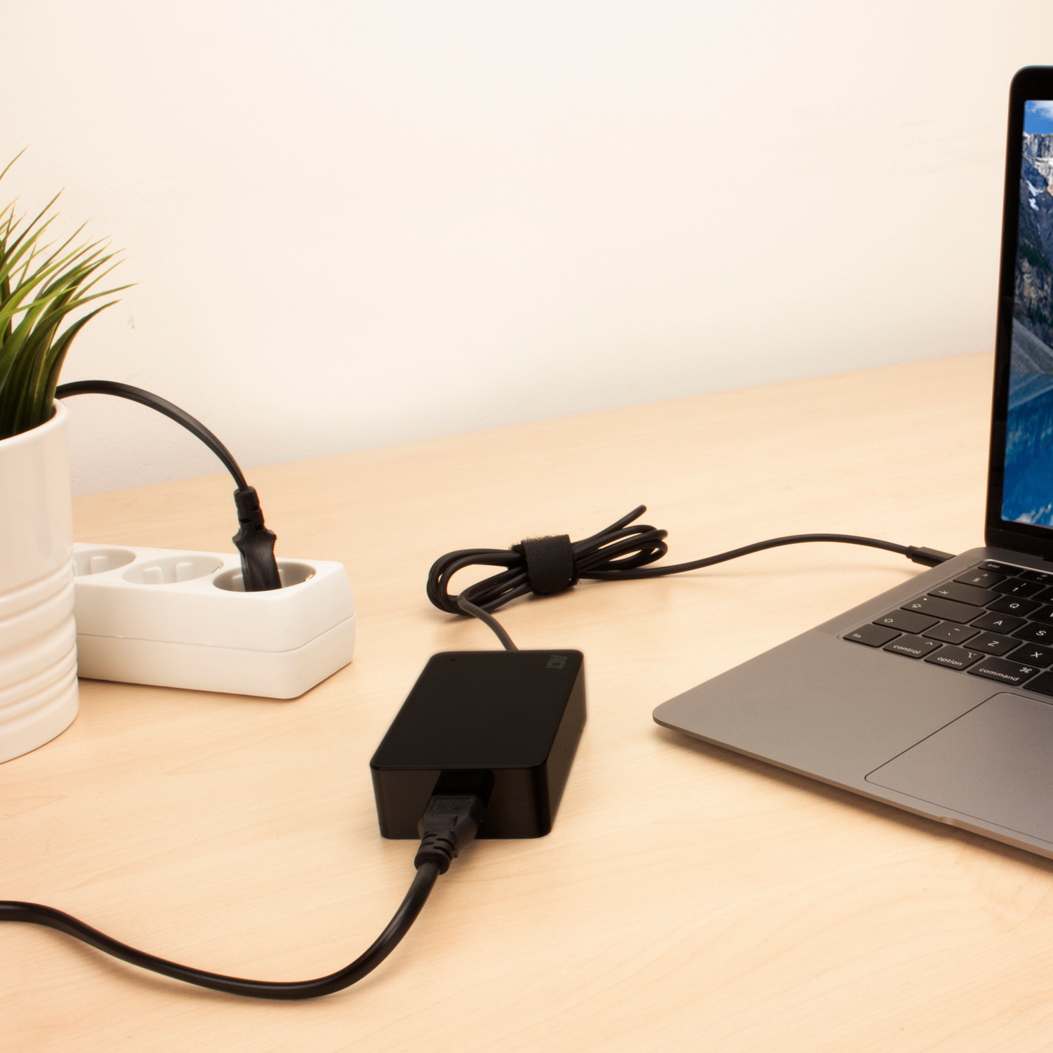Laptop connected to a black power adapter and a power strip on a wooden desk.
