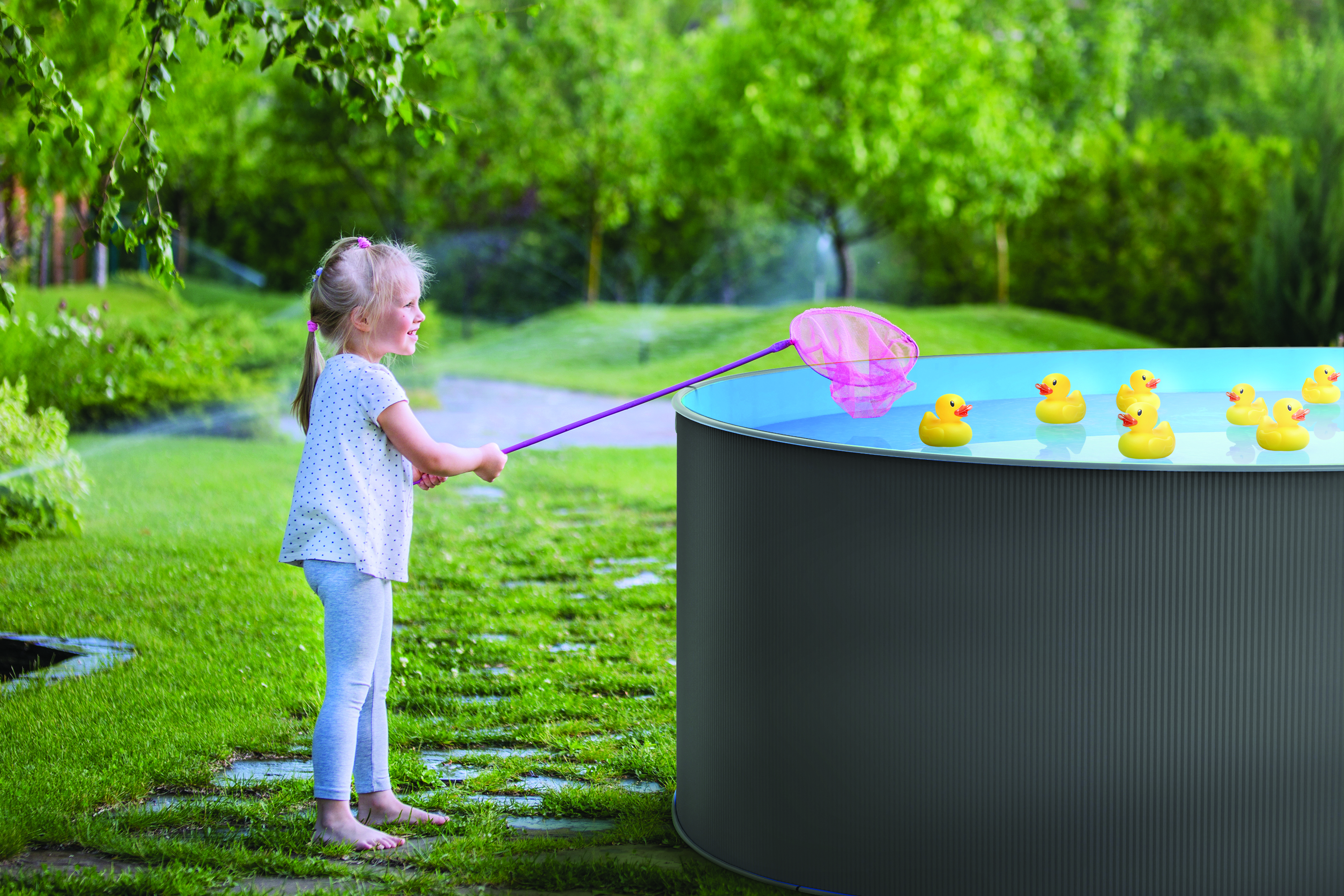 Mädchen, das ein Netz benutzt, um Gummienten in einem Pool zu fangen. Bäume und Gras im Hintergrund.