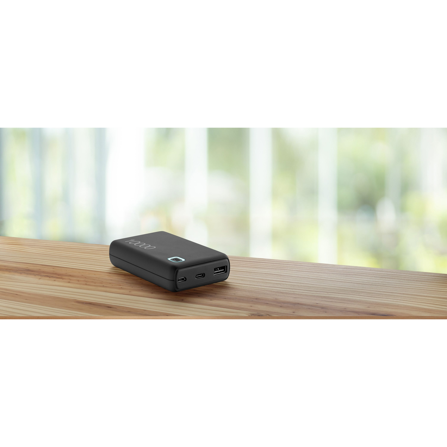 Schwarze Powerbank auf einem Holztisch. Der Hintergrund ist verschwommen, mit Fenster und Grün.