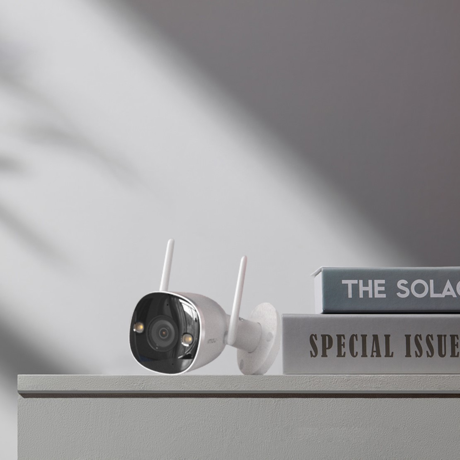 Biała kamera bezpieczeństwa z antenami, siedząca na szarej powierzchni, książki w tle.