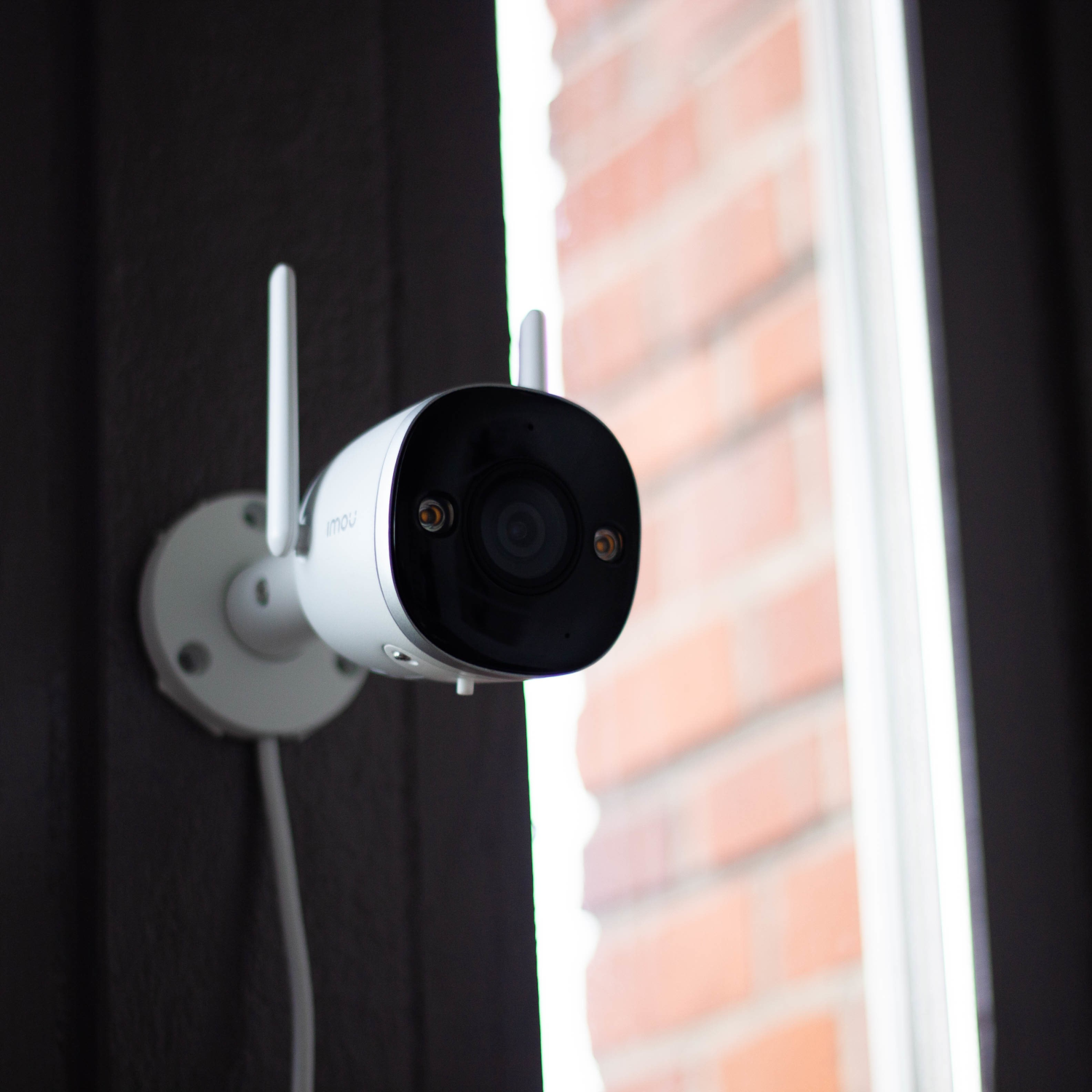 Biała kamera bezpieczeństwa zamontowana na ciemnej ścianie w pobliżu okna z ceglaną elewacją.