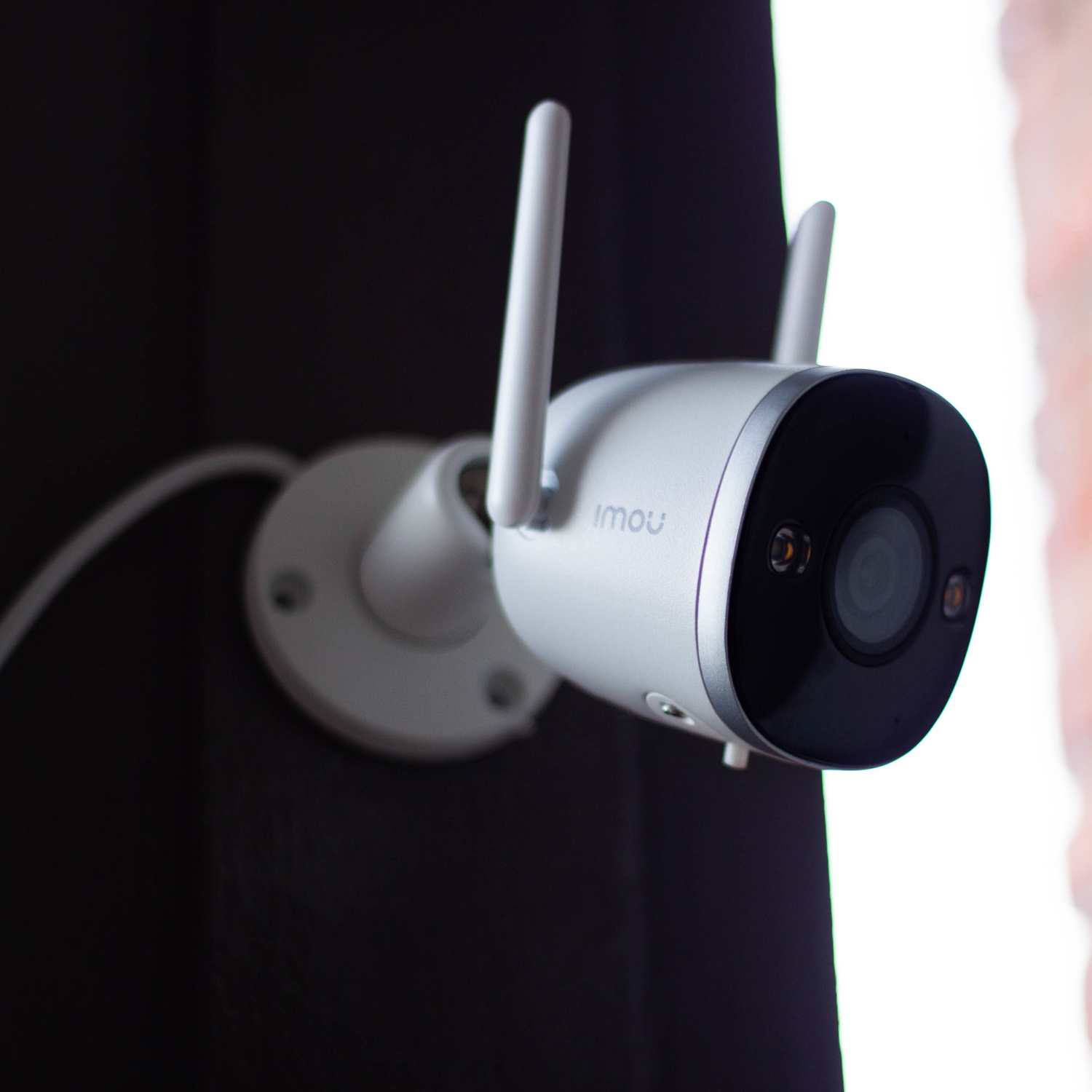 Biała kamera bezpieczeństwa Imou zamontowana na ciemnej powierzchni, z dwoma antenami, skierowana w prawo.