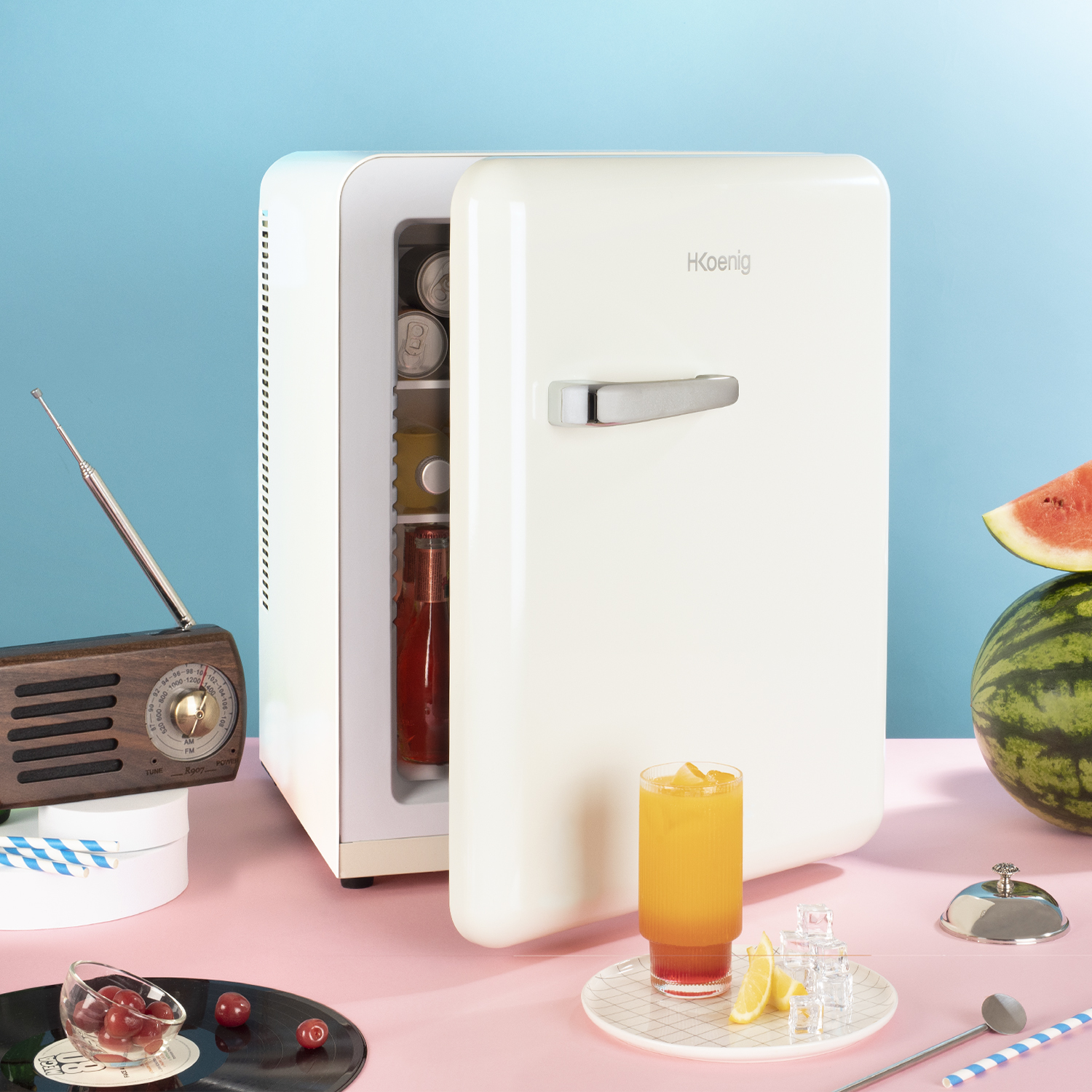 Un mini frigo retrò con la porta aperta, mostra bevande. Radio, decorazioni e fetta di anguria a fianco.