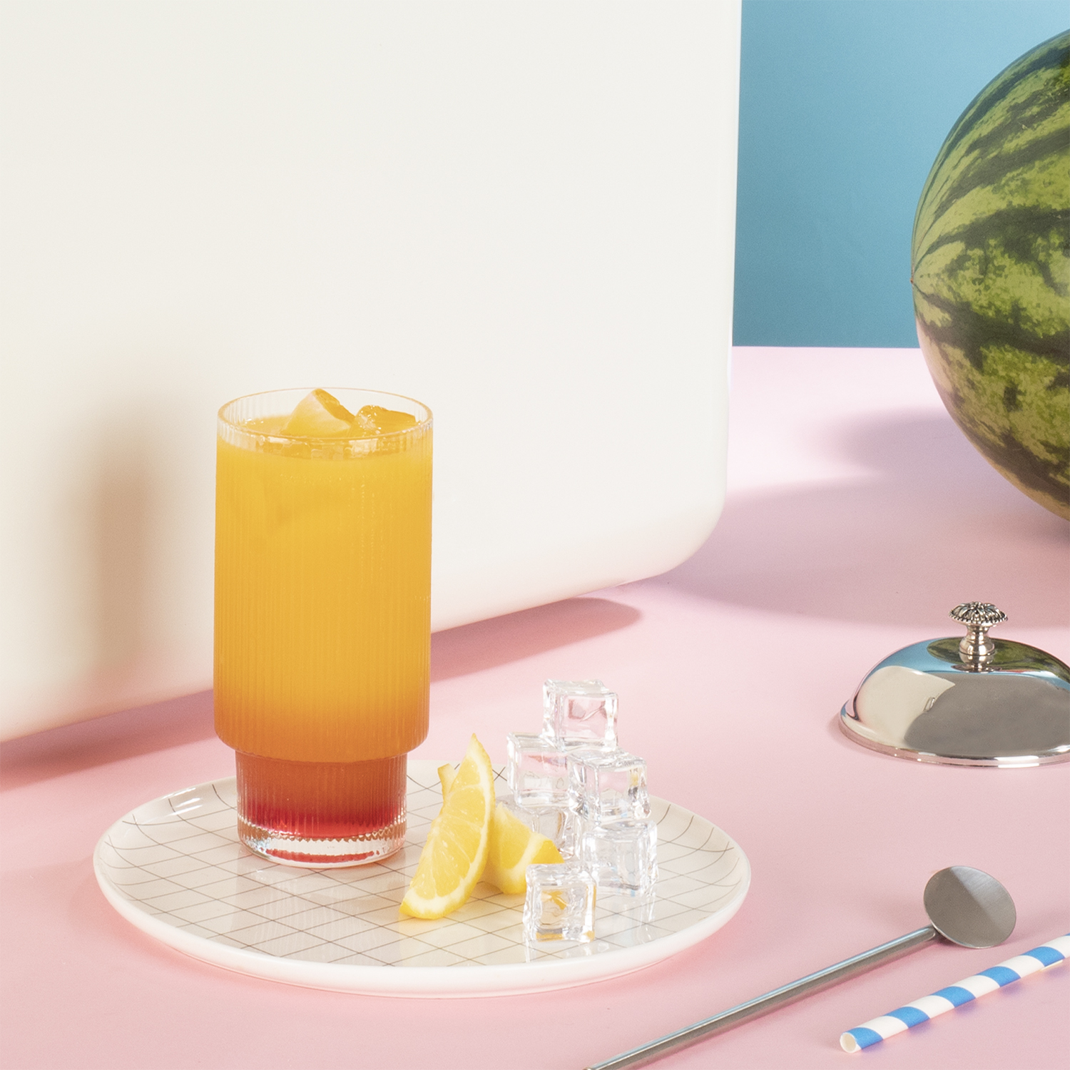 Bicchiere con bevanda arancione, ghiaccio e fette di limone su superficie rosa, sfondo bianco e blu.