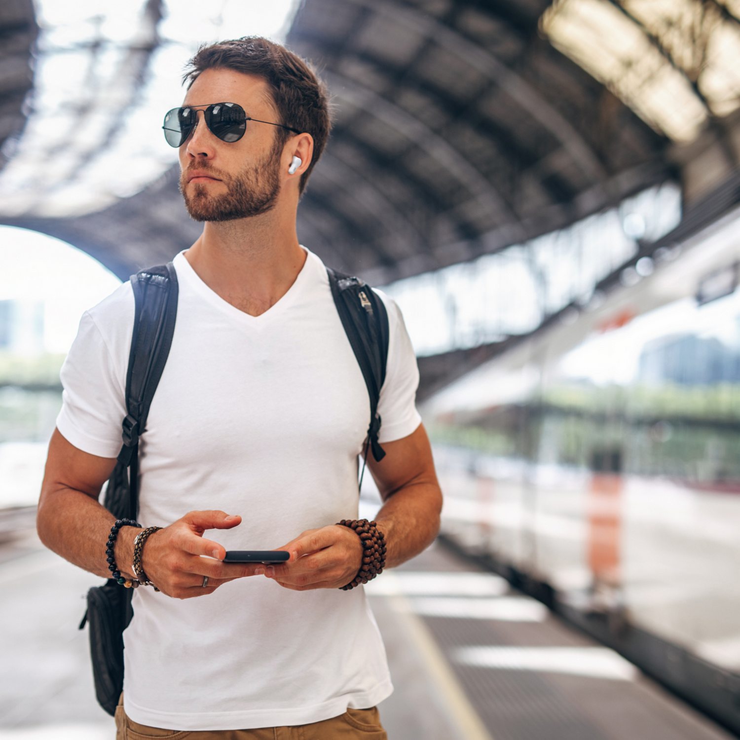 Hombre con gafas de sol y auriculares en una estación de tren, sosteniendo un teléfono.