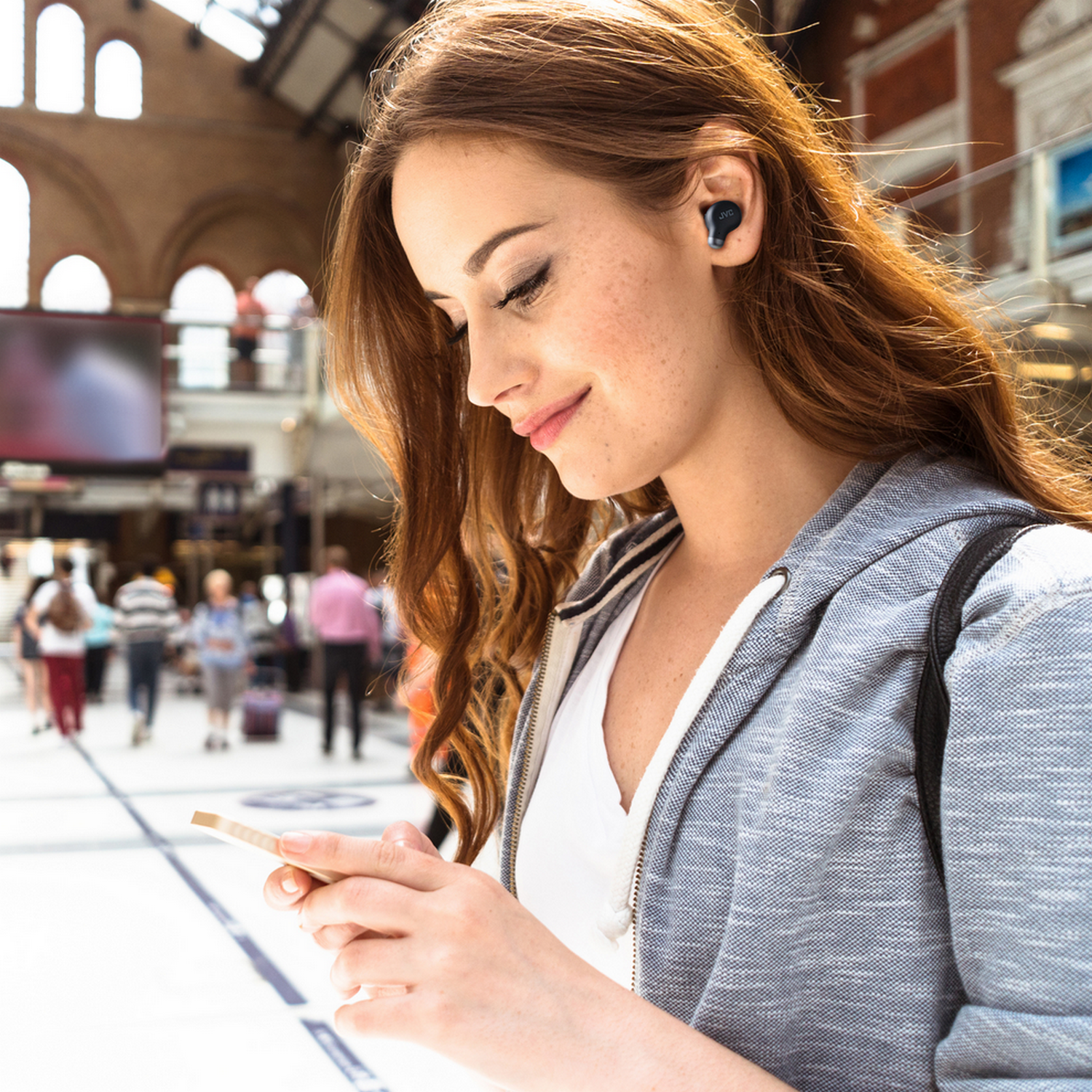 Eine Frau lächelt auf ein Telefon, trägt Ohrhörer, in einer geschäftigen Bahnhofsumgebung.