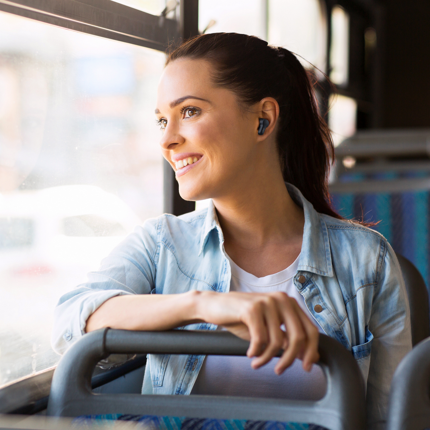 Kobieta w autobusie ze słuchawką, patrzy przez okno, uśmiecha się.